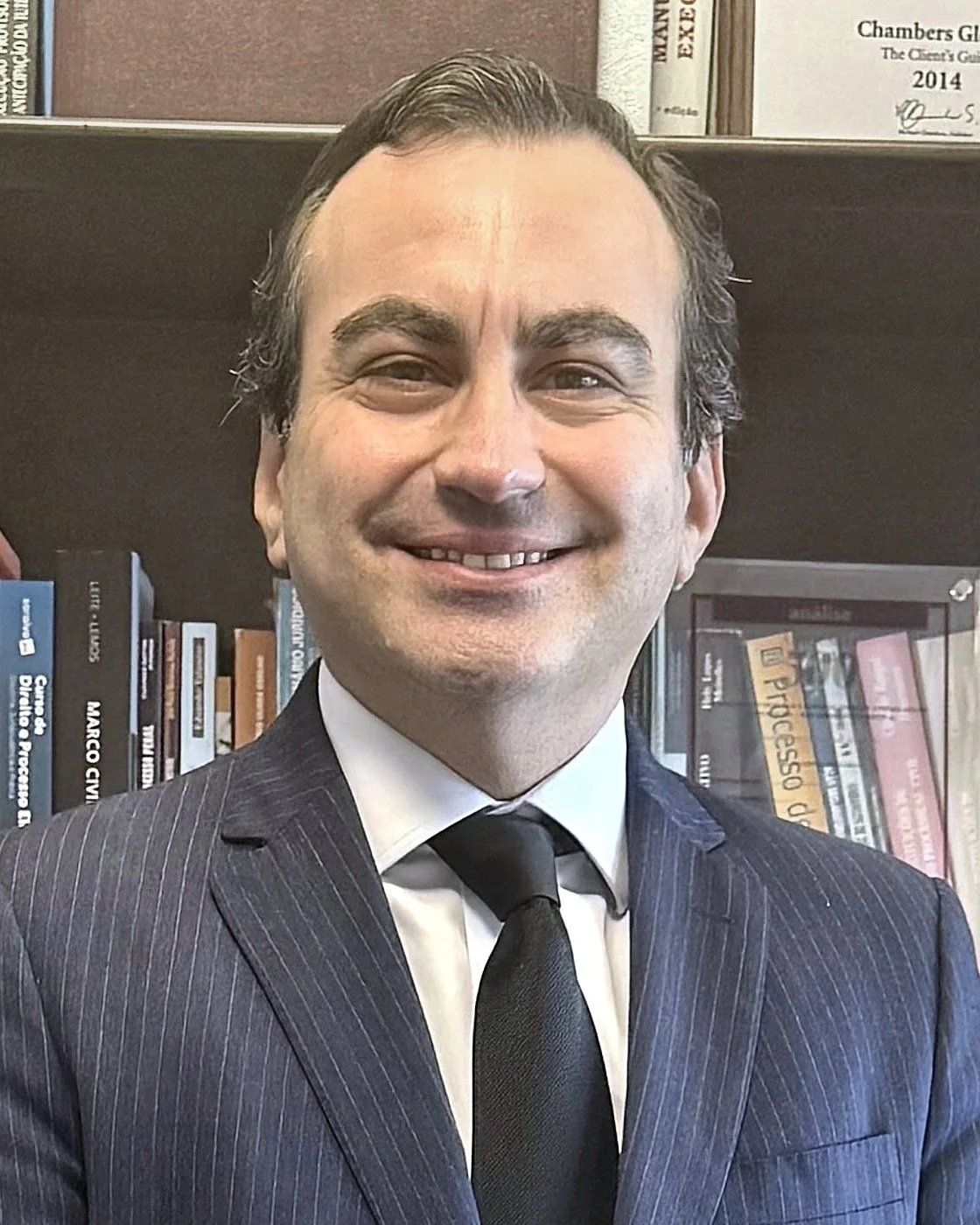A smiling man in a dark pinstripe suit, white shirt, and black tie, standing in front of a bookshelf filled with books and certificates.