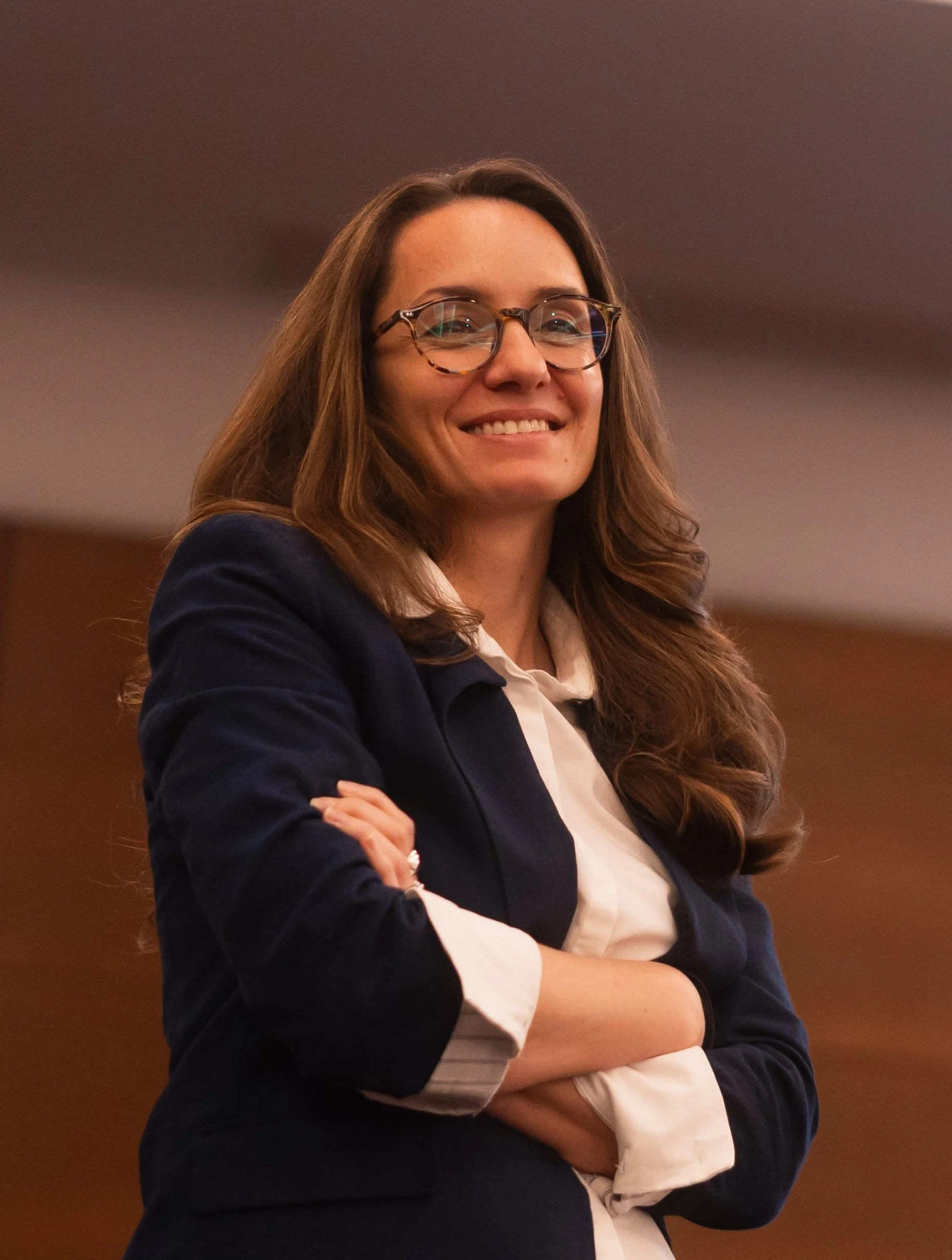 A woman with long wavy brown hair and glasses smiling, dressed in a white top and dark blazer, standing with crossed arms in an indoor setting.