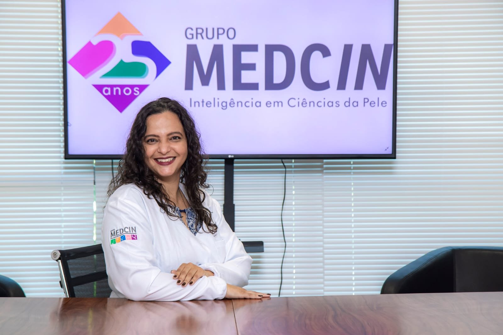 A woman with curly dark hair smiling at the camera, wearing a white lab coat with a colorful logo and the words 'Grupo MEDCIN' on the sleeve, sitting at a conference table in a room with window blinds and a monitor displaying the 'Grupo MEDCIN' logo and text.
