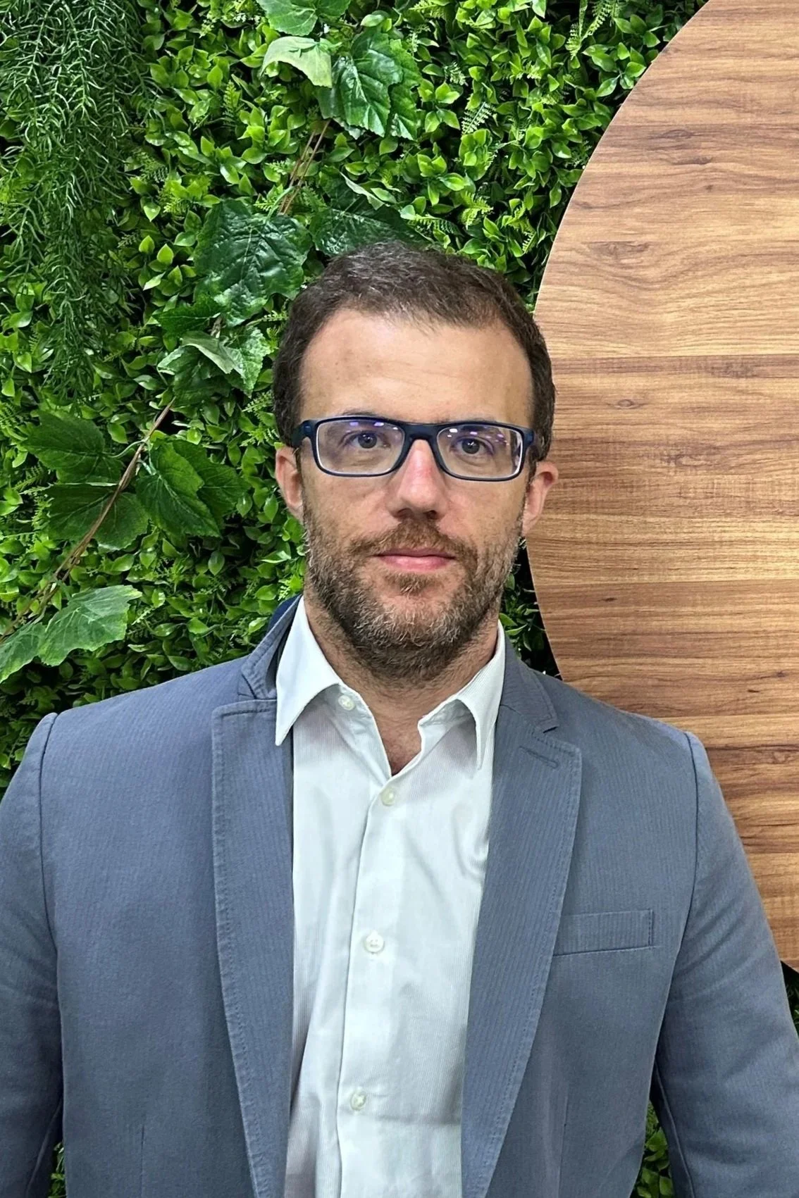 A man with glasses and a beard wearing a light blue suit and white dress shirt. He is standing in front of a green plant wall and wooden wall.