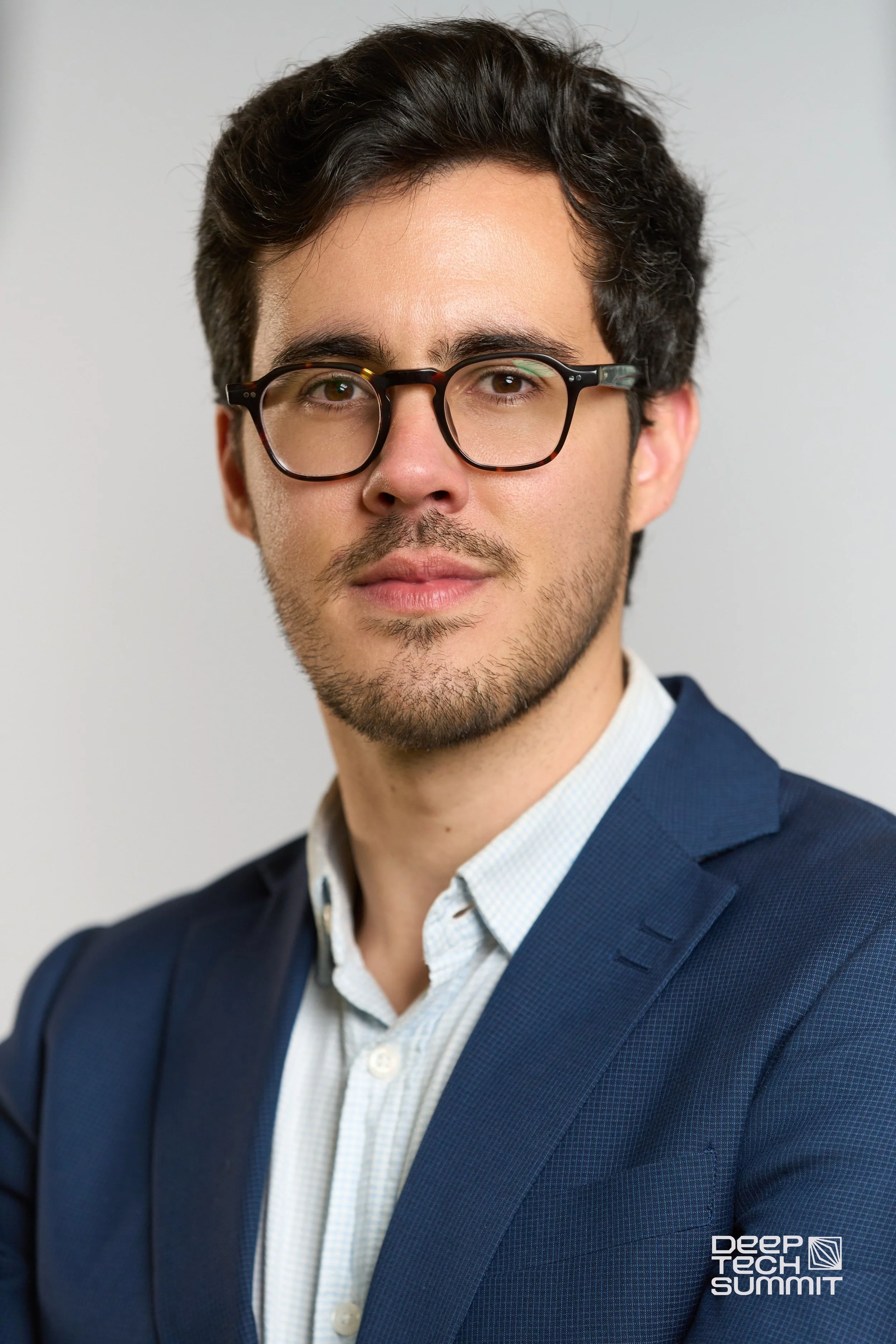 Close-up of a young man with dark hair, wearing glasses and a navy blue suit, at the Deep Tech Summit event.