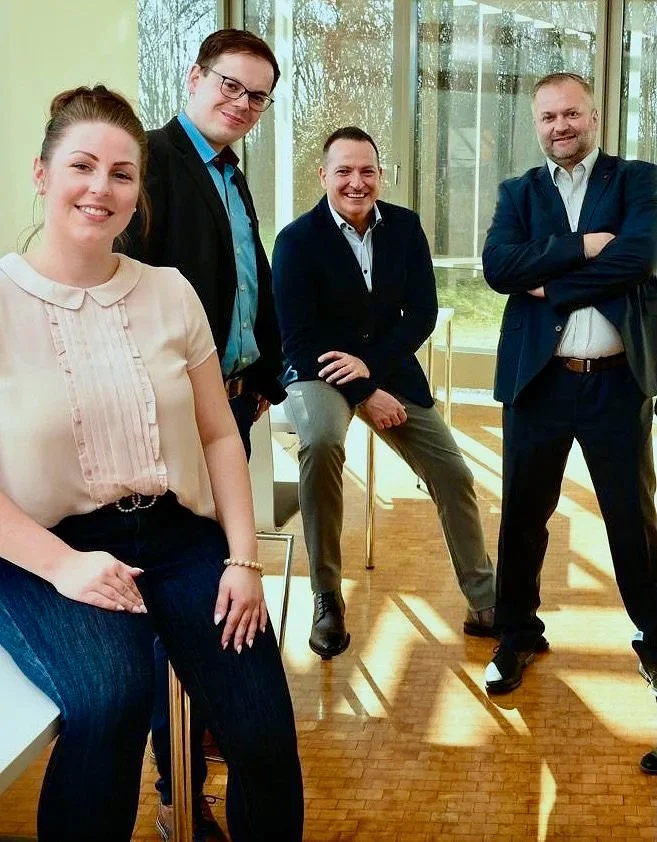 Vier Menschen, zwei Männer und zwei Frauen, stehen im Raum vor großen Fenstern, lachen und posieren für die Kamera, im Hintergrund eine grüne Außenansicht.