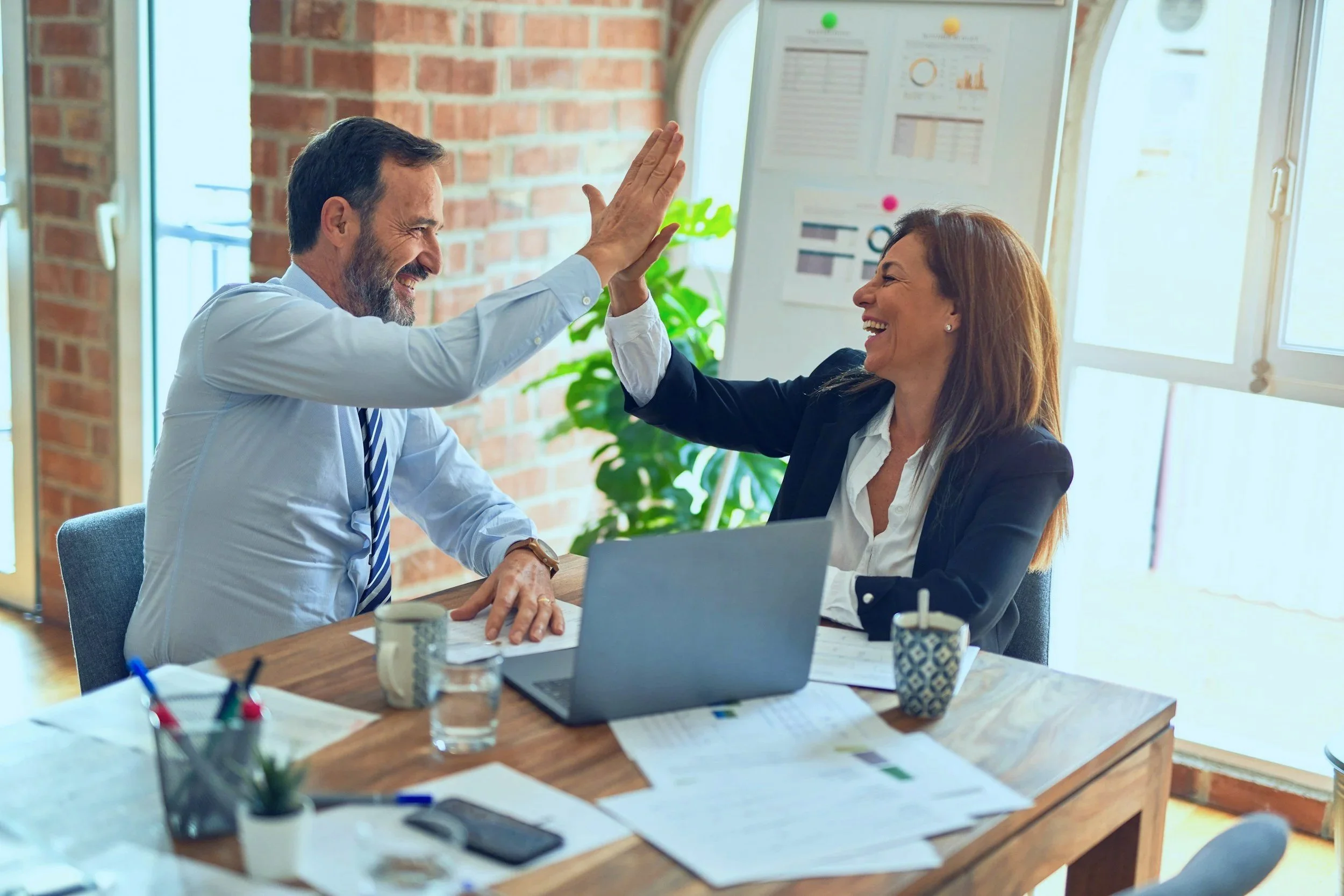 Ein Mann und eine Frau in einem Büro sitzen sich gegenüber und geben sich eine High-Five, während sie lachen.
