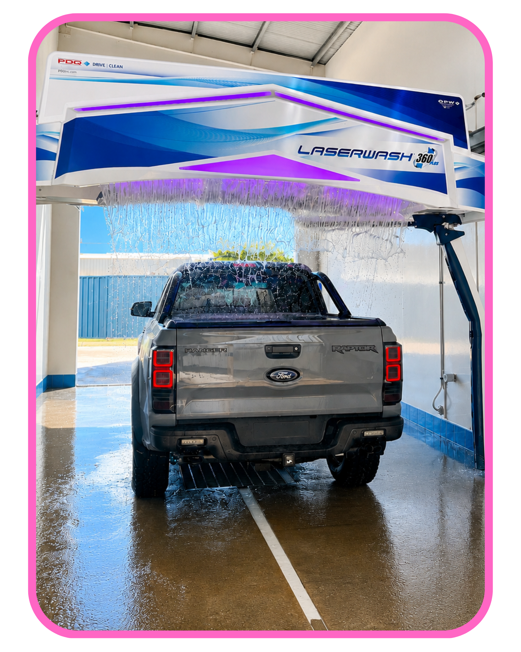 A silver Ford Ranger Raptor pickup truck is being washed under a car wash arch with purple and blue lights. Water is flowing over the vehicle, and the floor is wet.