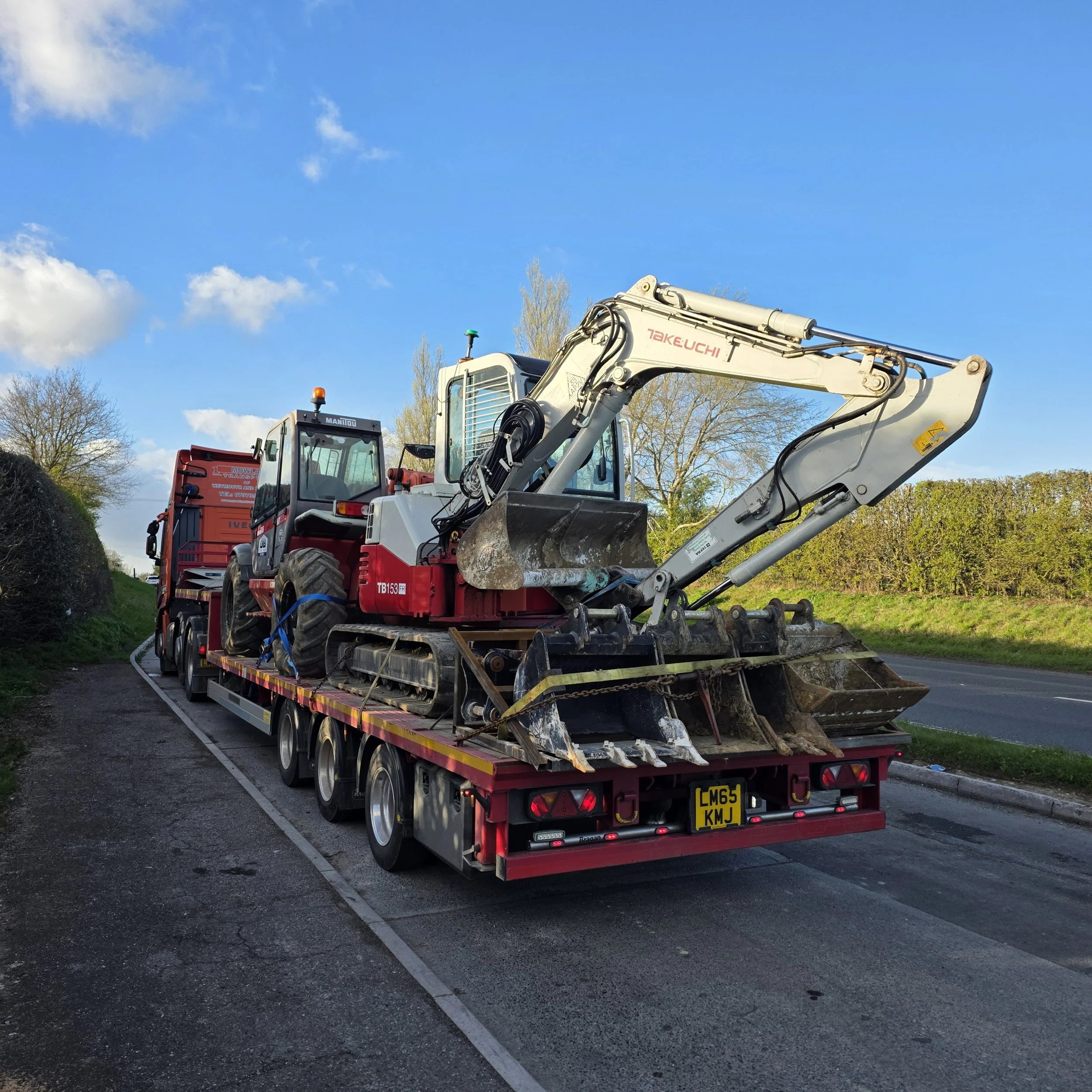 Construction excavator on a flatbed truck on a paved road surrounded by trees and blue sky.
