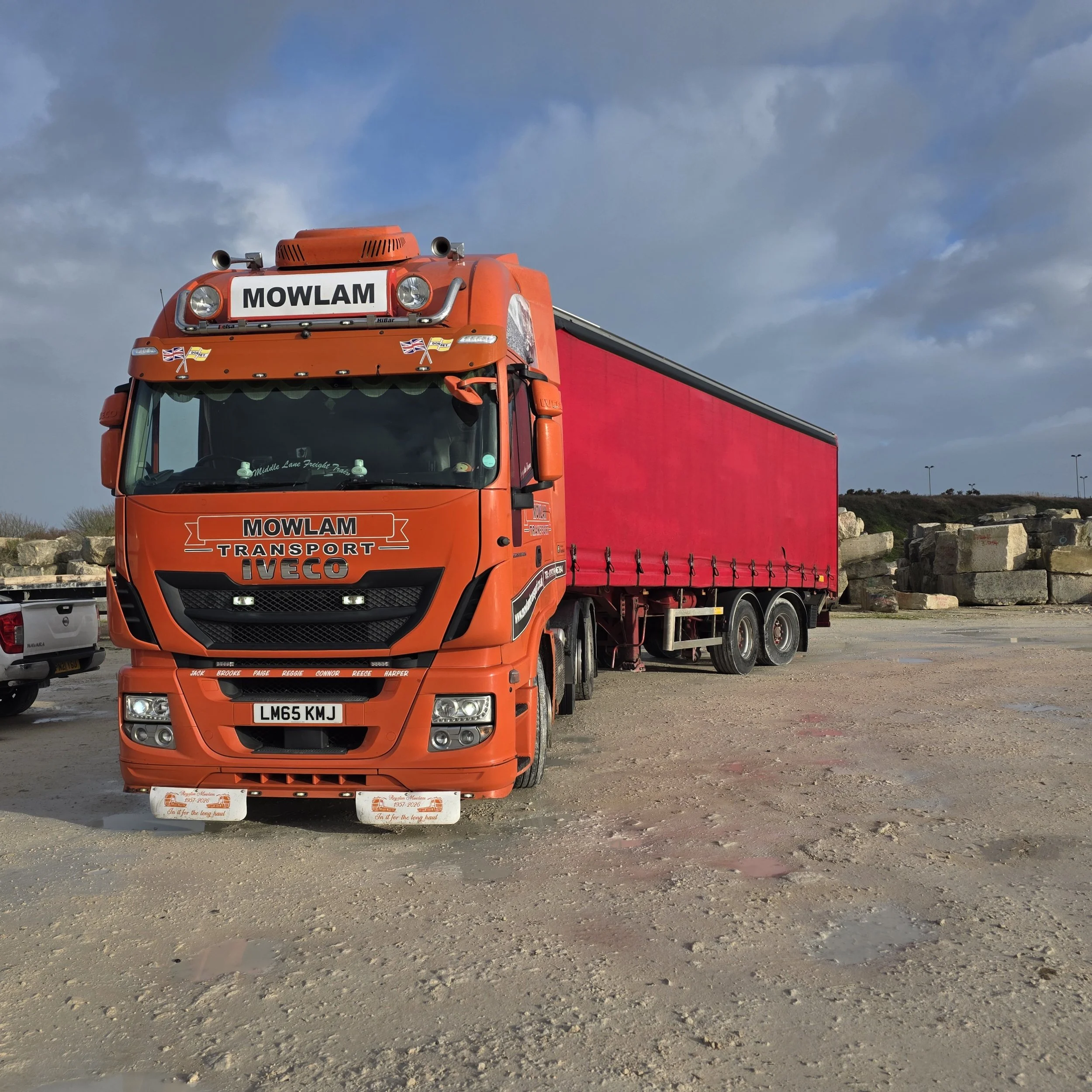 A bright orange freight truck with a red trailer parked on a gravel lot under a partly cloudy sky.