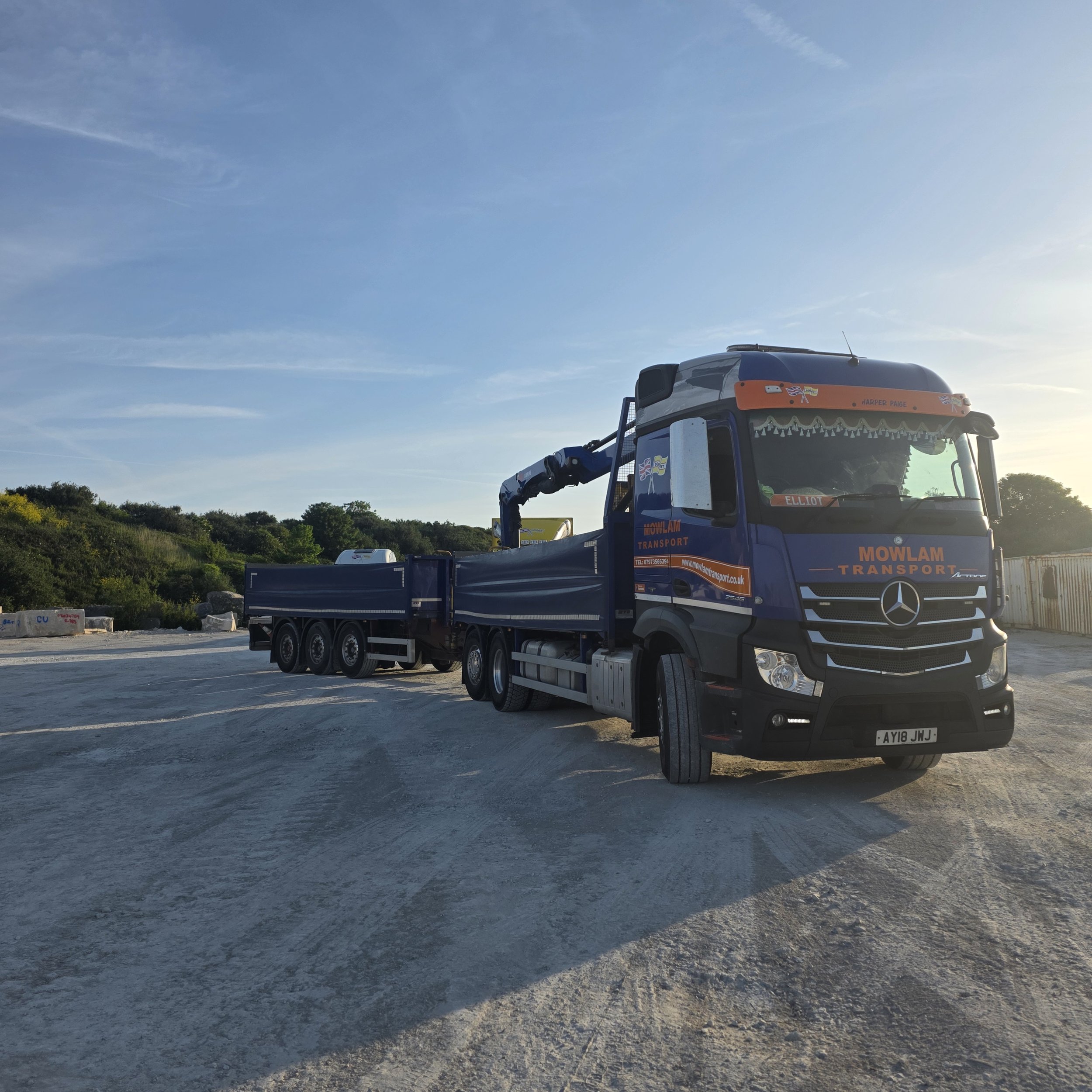 A large truck with a blue and orange color scheme, labeled 'Mowlam Transport,' parked outdoors under a clear sky with sparse clouds. Trees are visible in the background.