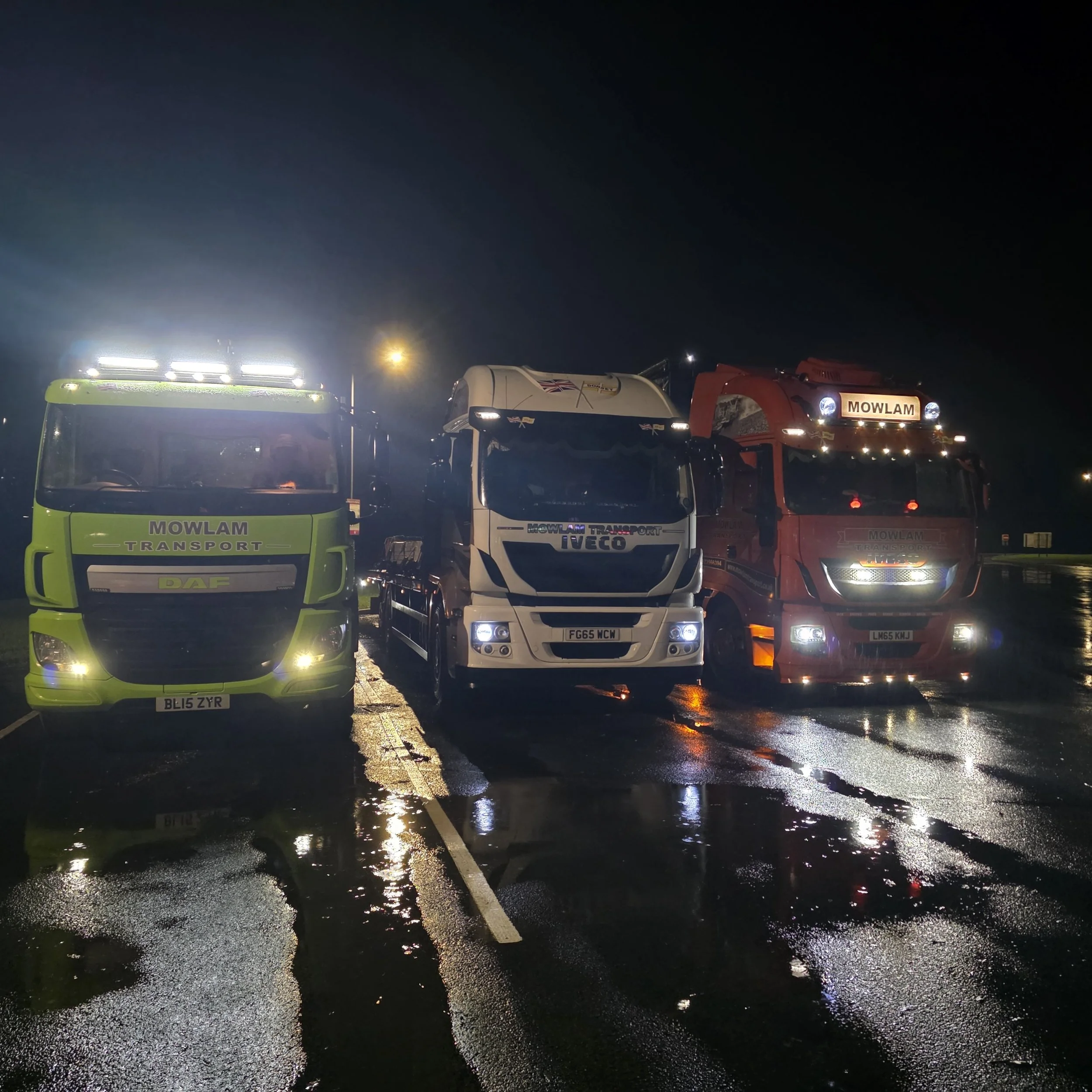Nighttime parking lot scene with several trucks, their lights reflected on wet pavement, including a green truck on the left, a white truck in the center, and a red truck on the right, with a dark sky in the background.