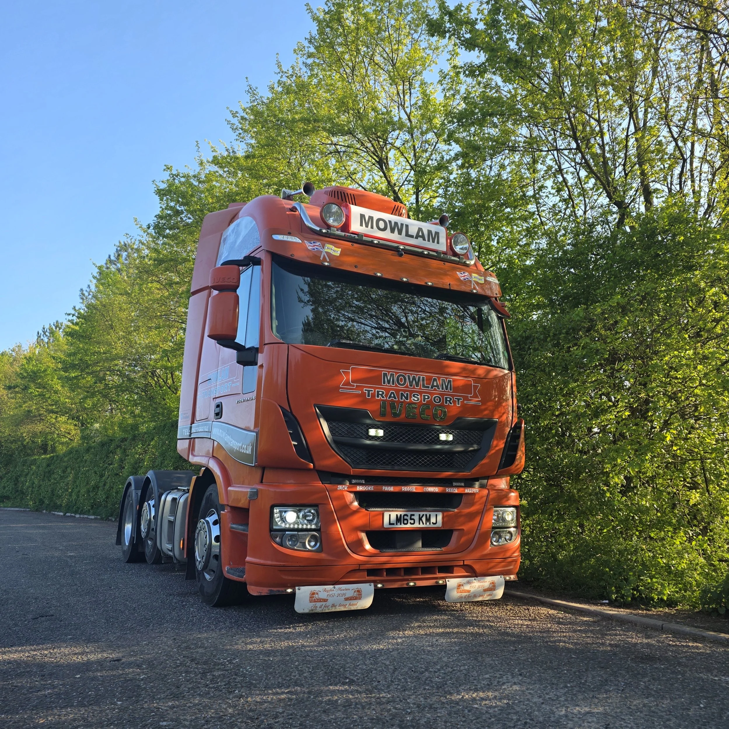 Red transport truck with MOWLAM TRANSORT logo, parked under trees with green leaves and a blue sky.