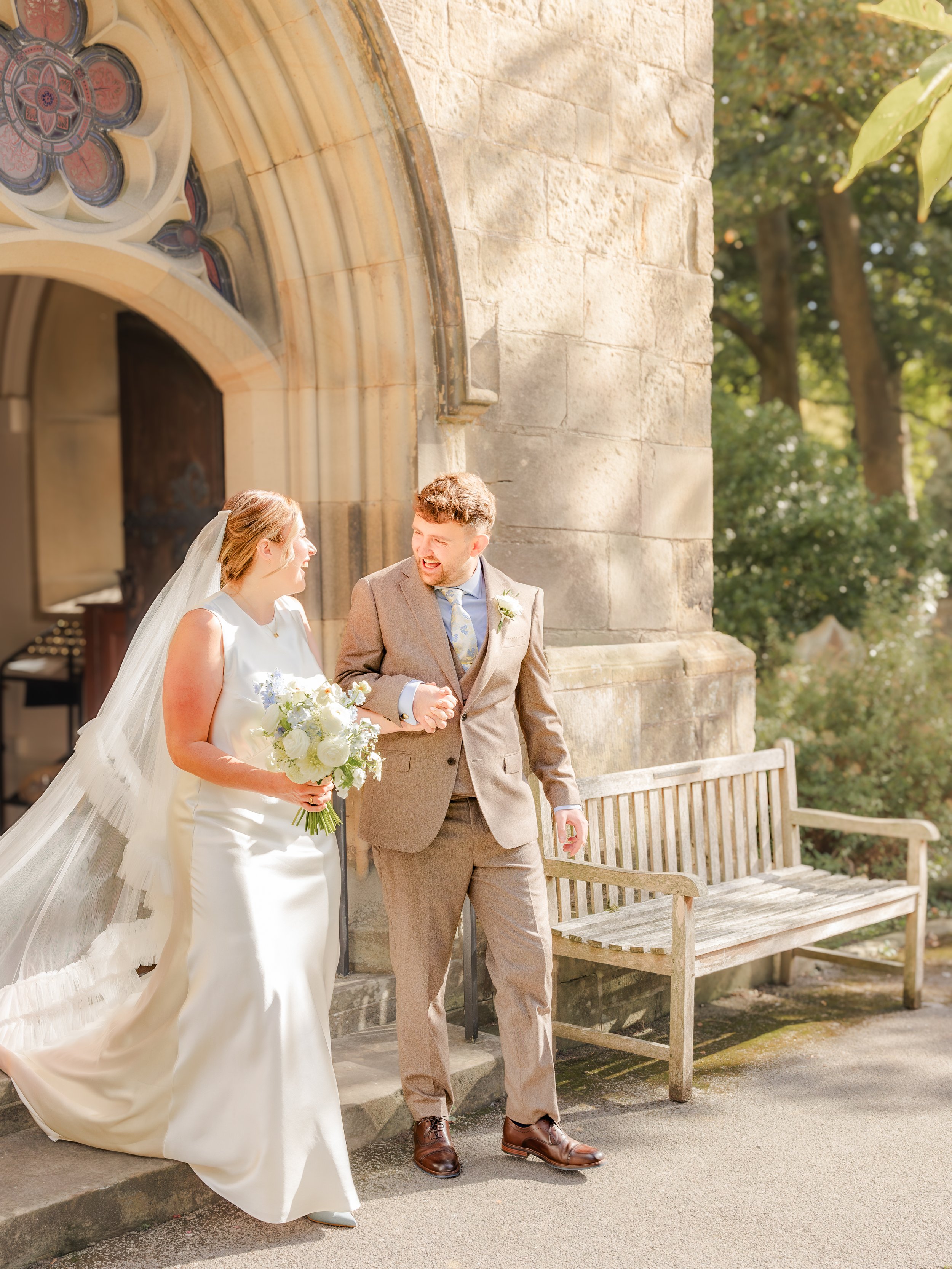 A bride and groom walking and smiling outside a church with stone walls and a wooden bench.