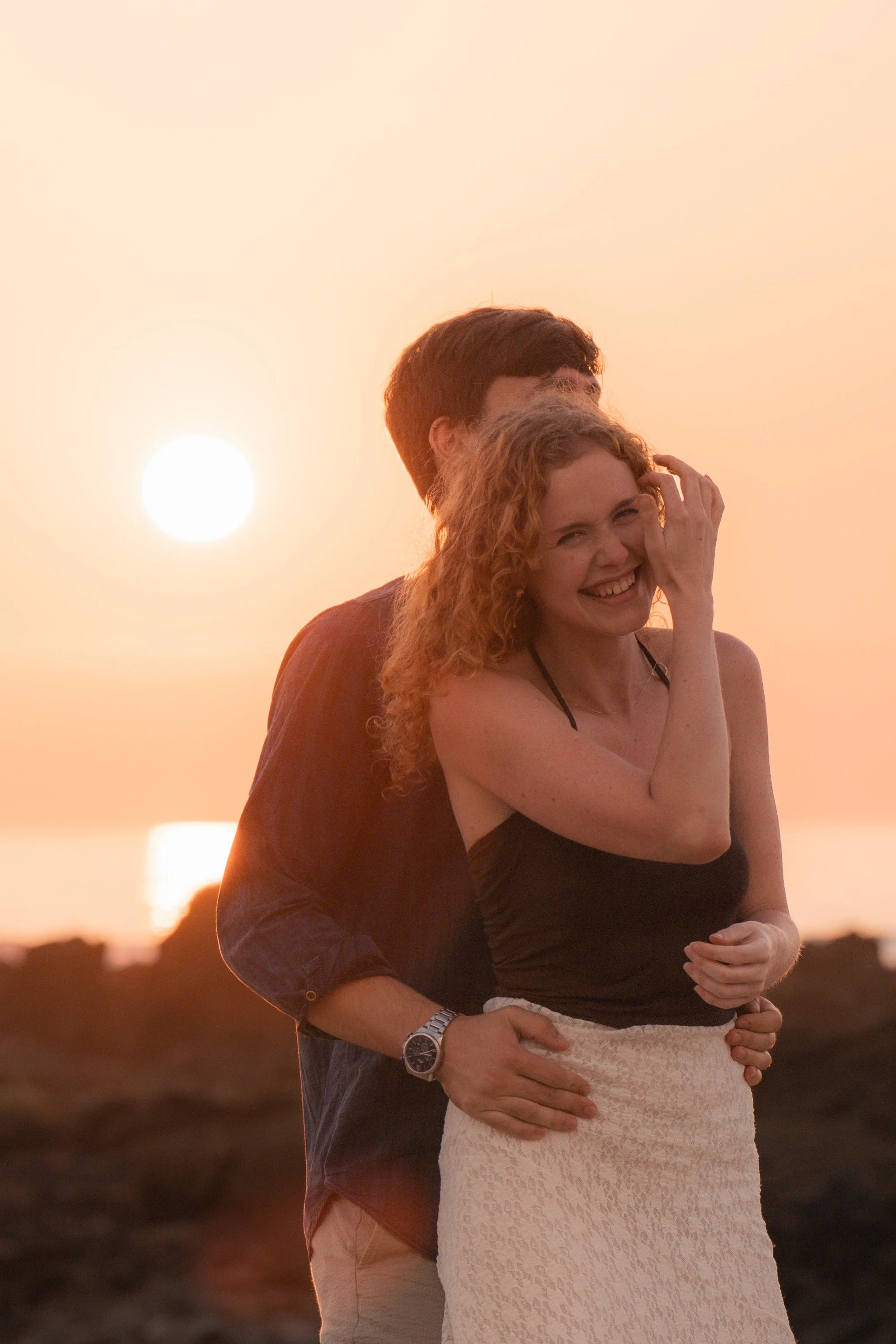 A couple embracing each other at sunset, with the woman smiling and touching her face, while the man is hugging her from behind. The sunset creates a warm glow in the background.