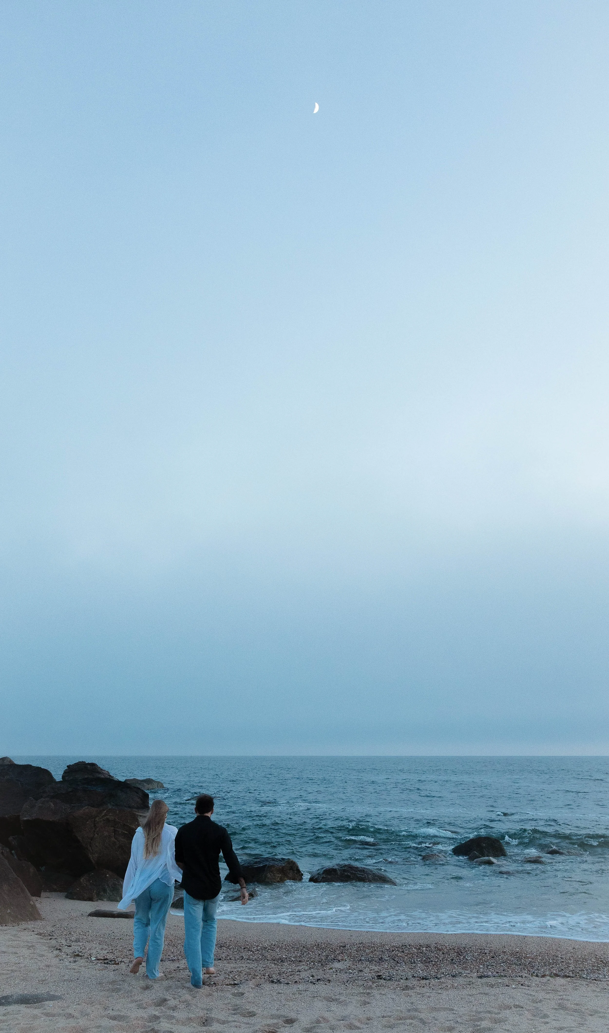 A man and a woman walking on a beach near rocks, with the ocean and a clear sky with a crescent moon in the background.