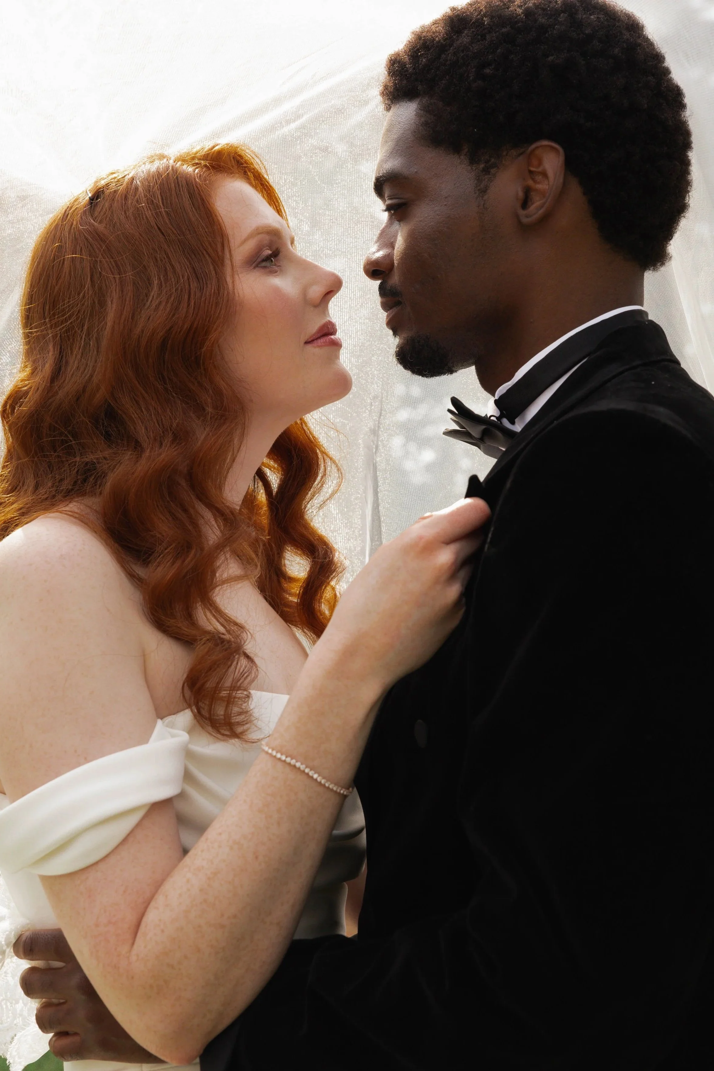 A bride and groom face closely, gazing into each other's eyes during a wedding. The bride is a red-haired woman wearing a white off-shoulder dress and a pearl bracelet, holding the groom's shoulder. The groom is a dark-haired man dressed in a tuxedo with a bow tie, gently touching the bride.