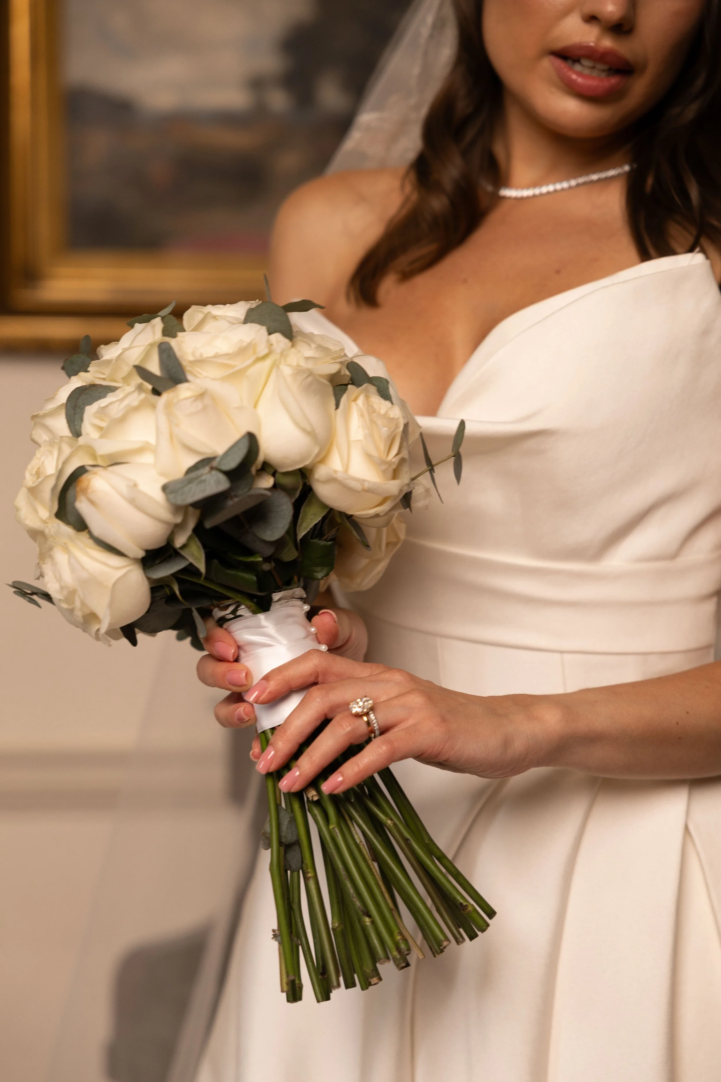 A woman dressed in a white wedding gown holding a bouquet of white roses and greenery, wearing a pearl necklace, with her lips slightly parted, in an indoor setting.