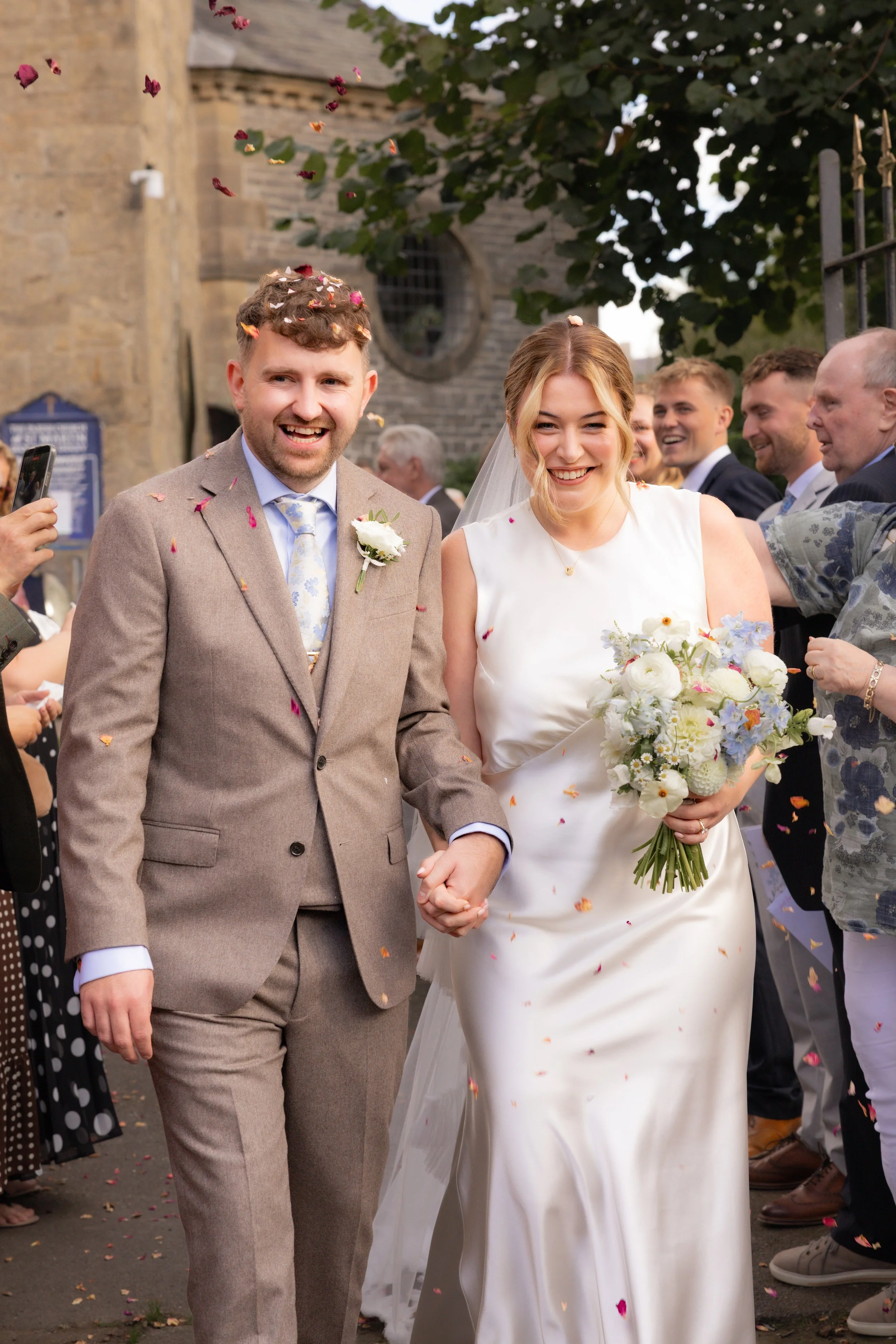 A bride and groom smiling and holding hands during their wedding celebration, surrounded by guests throwing flower petals.