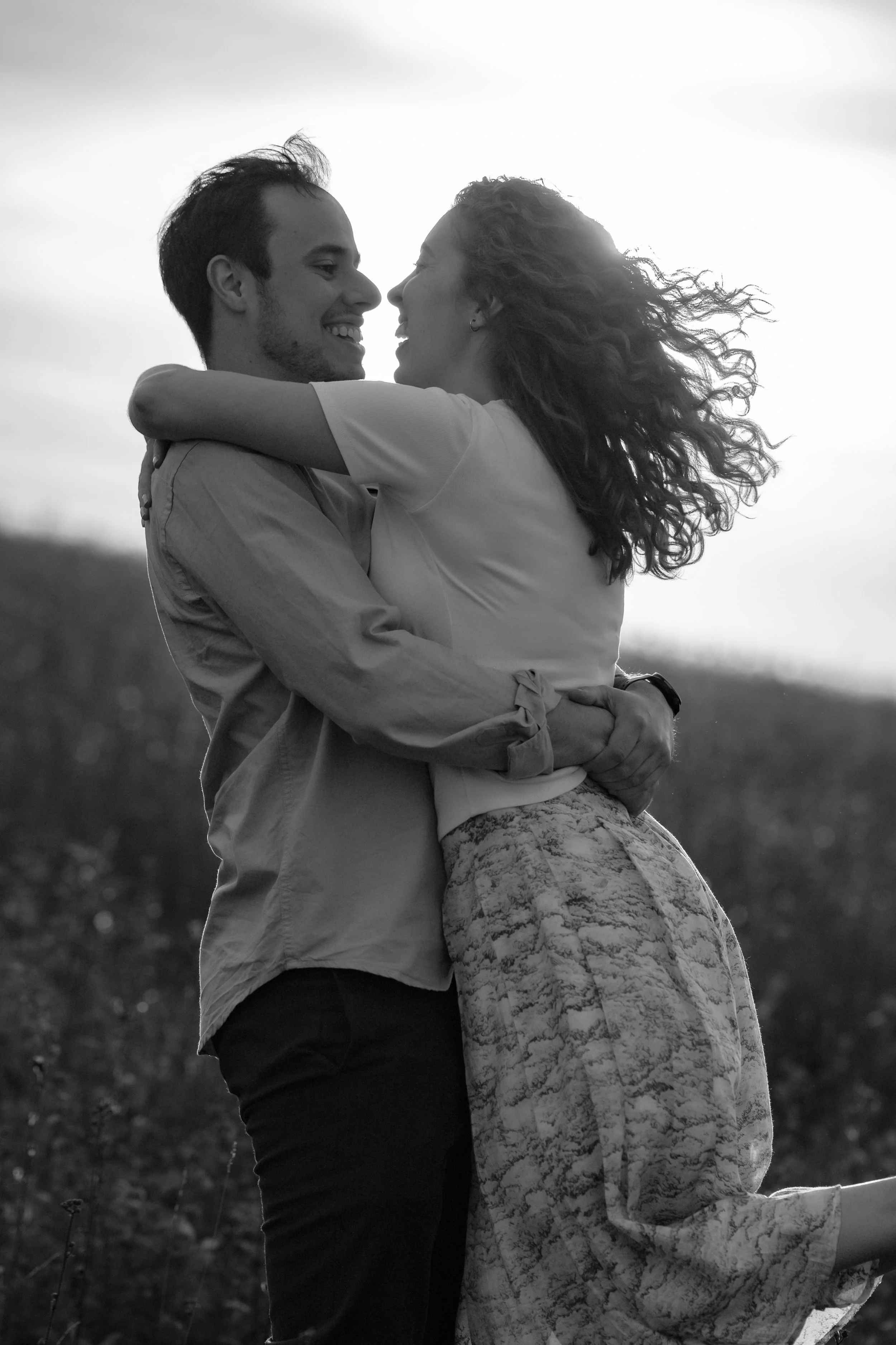 A black-and-white photo of a couple embracing outdoors, smiling and looking into each other's eyes, with a blurred natural background.
