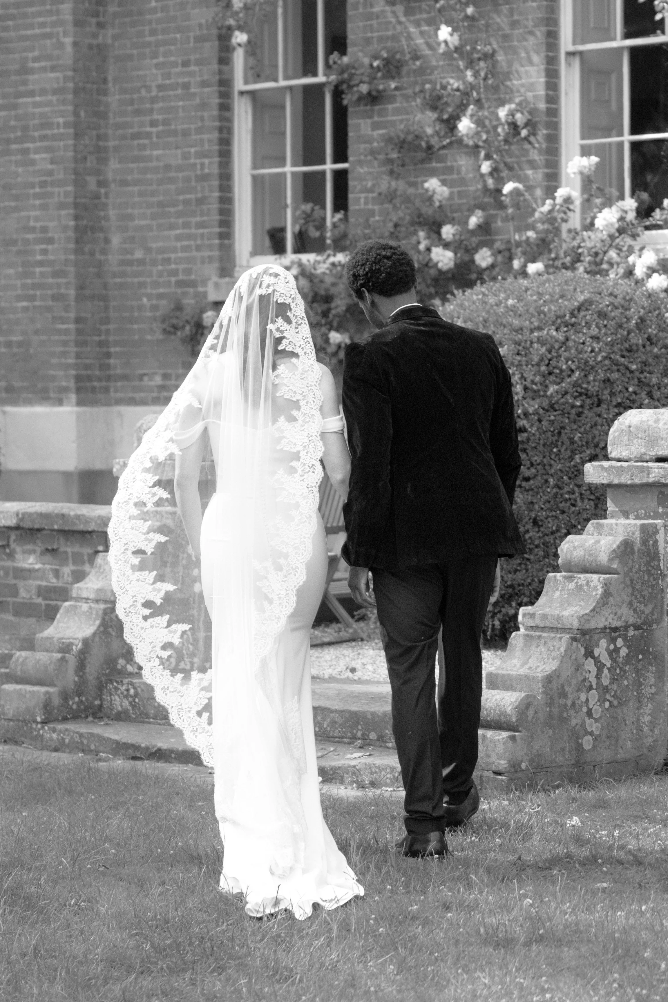 A black-and-white photo of a bride and groom walking away from the camera on stairs outside a brick house with blooming bushes. The bride wears a wedding dress and veil, the groom wears a dark suit.