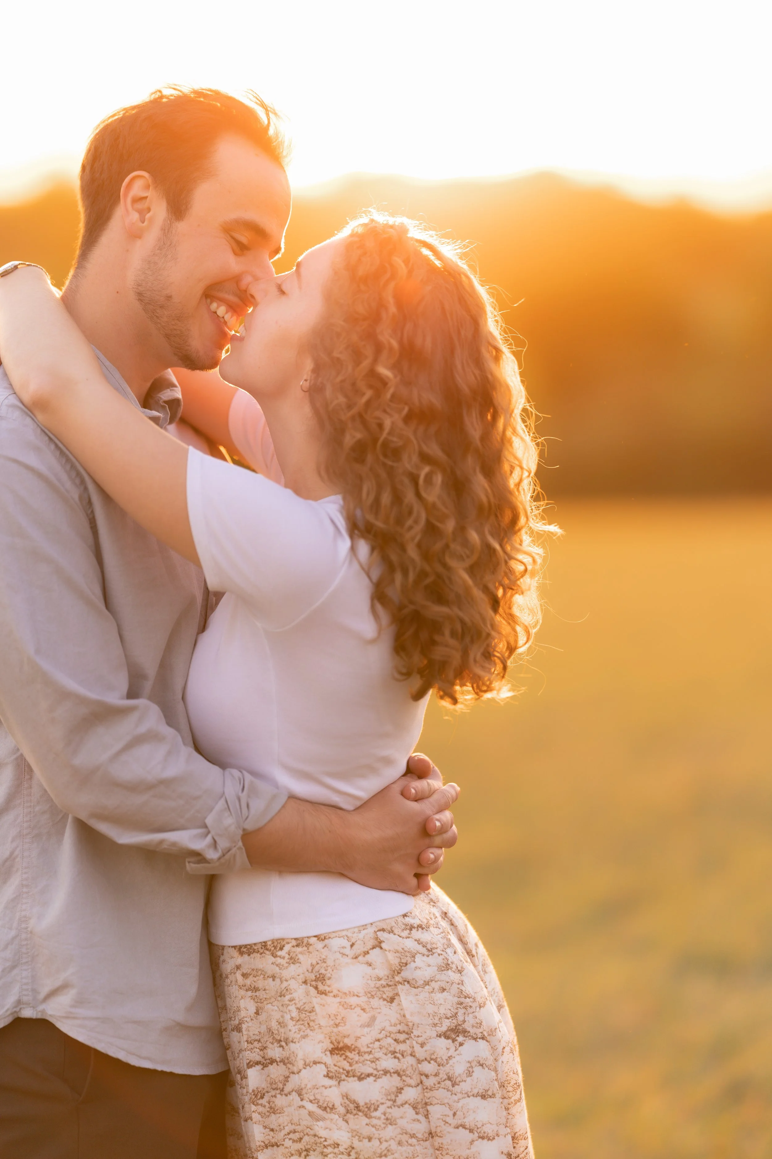 A couple embraces and shares a gentle kiss outdoors during sunset, with the sun setting behind them and a blurred landscape in the background.