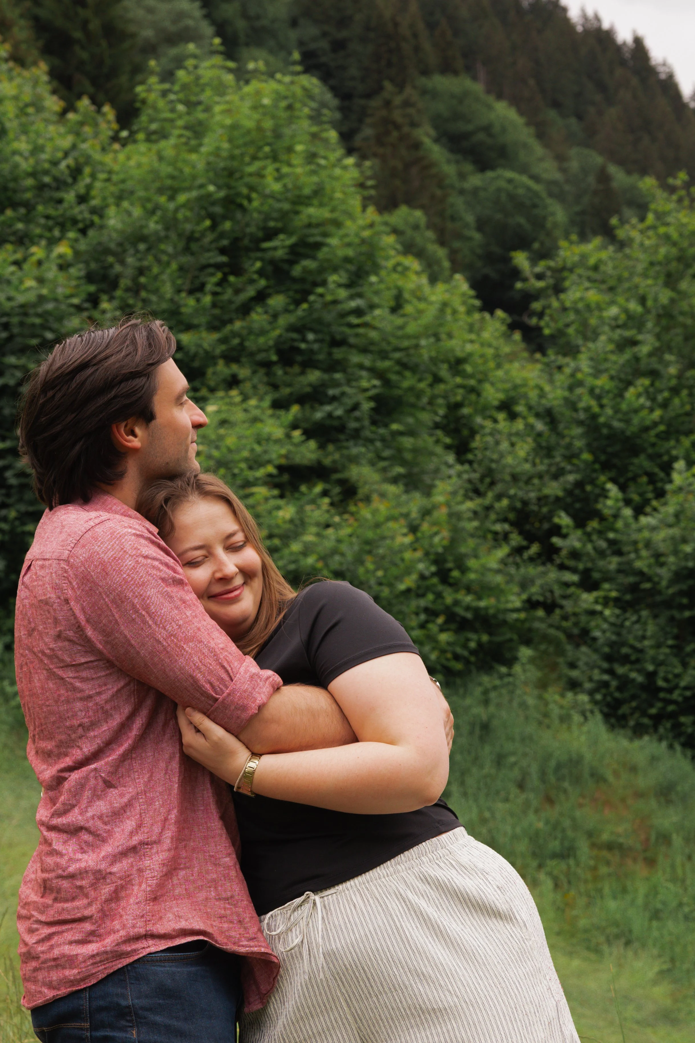 A man and woman hugging outdoors in front of green trees and grass, both smiling with eyes closed.