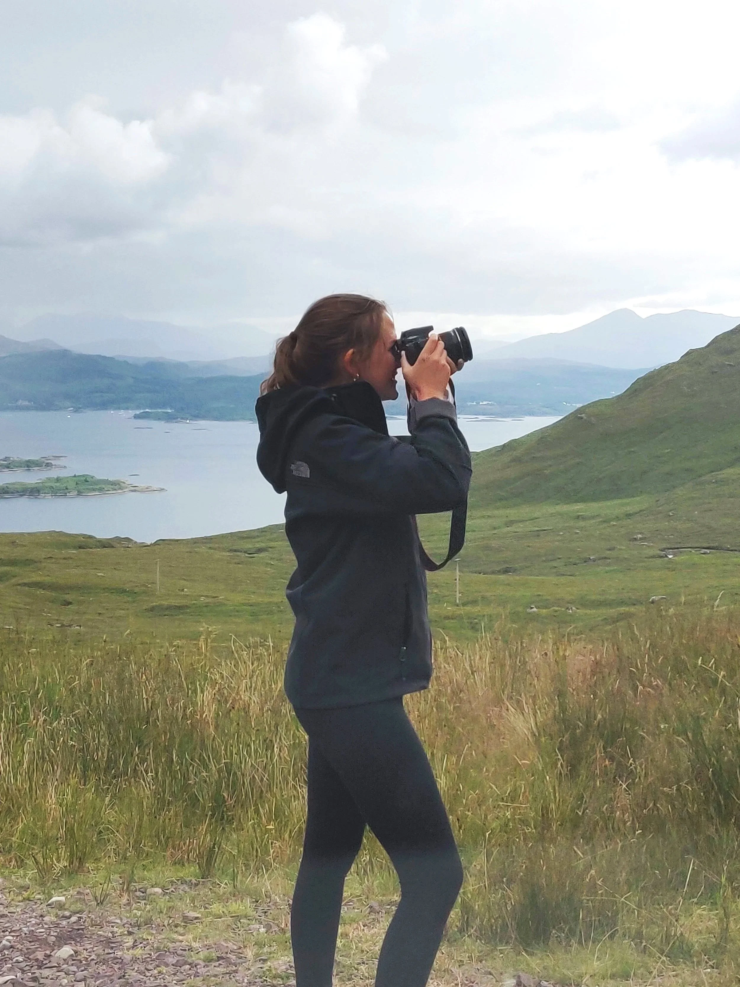A woman with brown hair tied in a ponytail takes a photograph with a camera in a grassy, mountainous landscape during daytime.