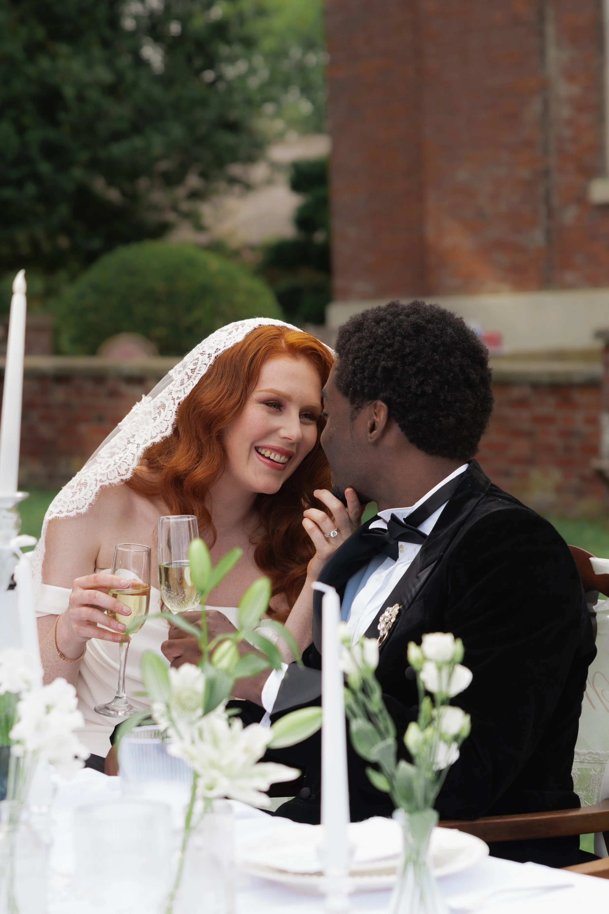 A bride with long red hair wearing a lace veil and a white wedding dress, smiling while holding a glass of champagne. She is sitting outdoors with a groom who has short curly black hair, wearing a black tuxedo with a bow tie. They are at a wedding reception table decorated with white flowers and green foliage, with a brick building and green trees in the background.