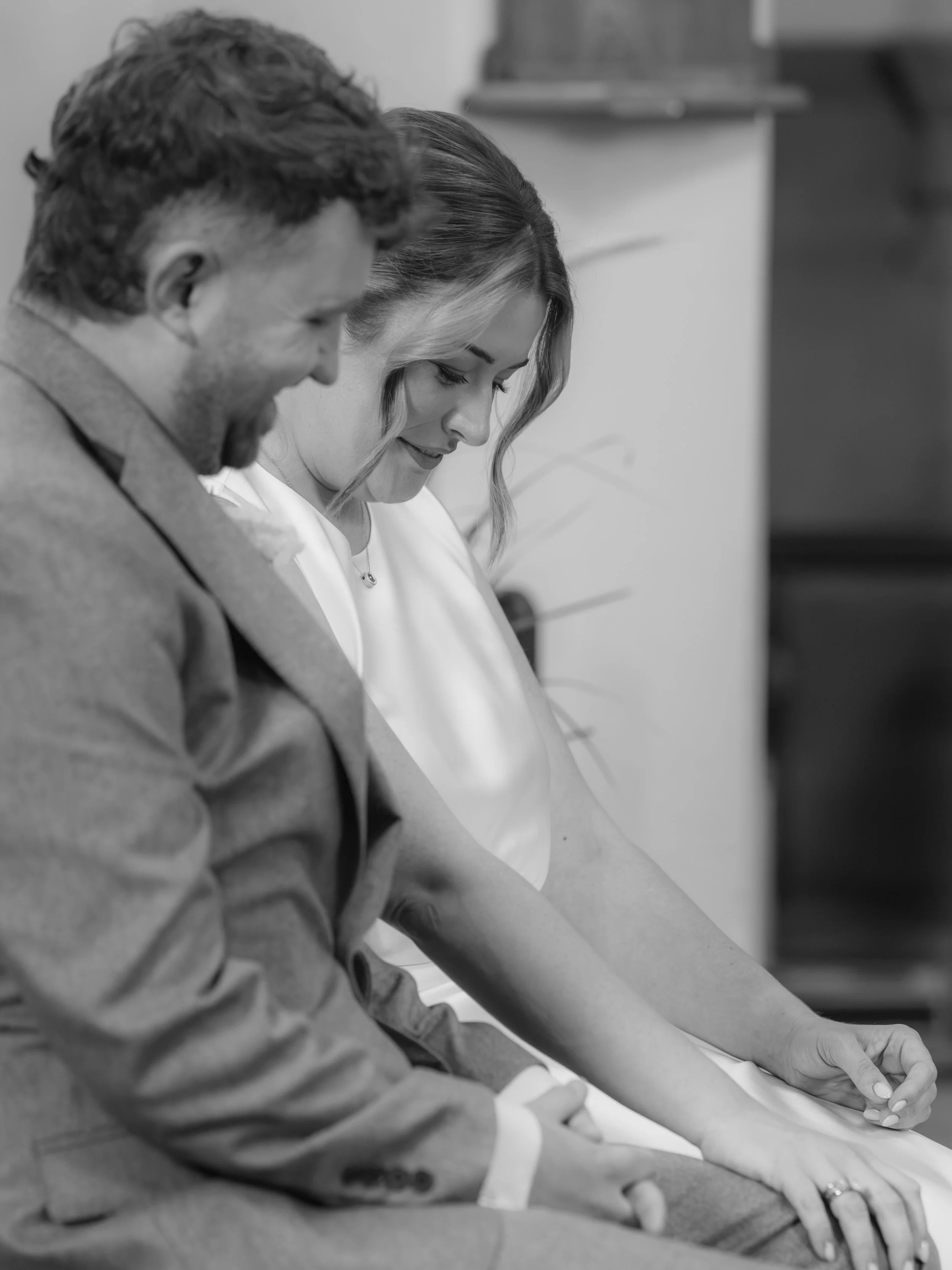 Black and white photo of two people sitting at their ceremony looking down, with focus on their hands and faces, leaning towards each other.