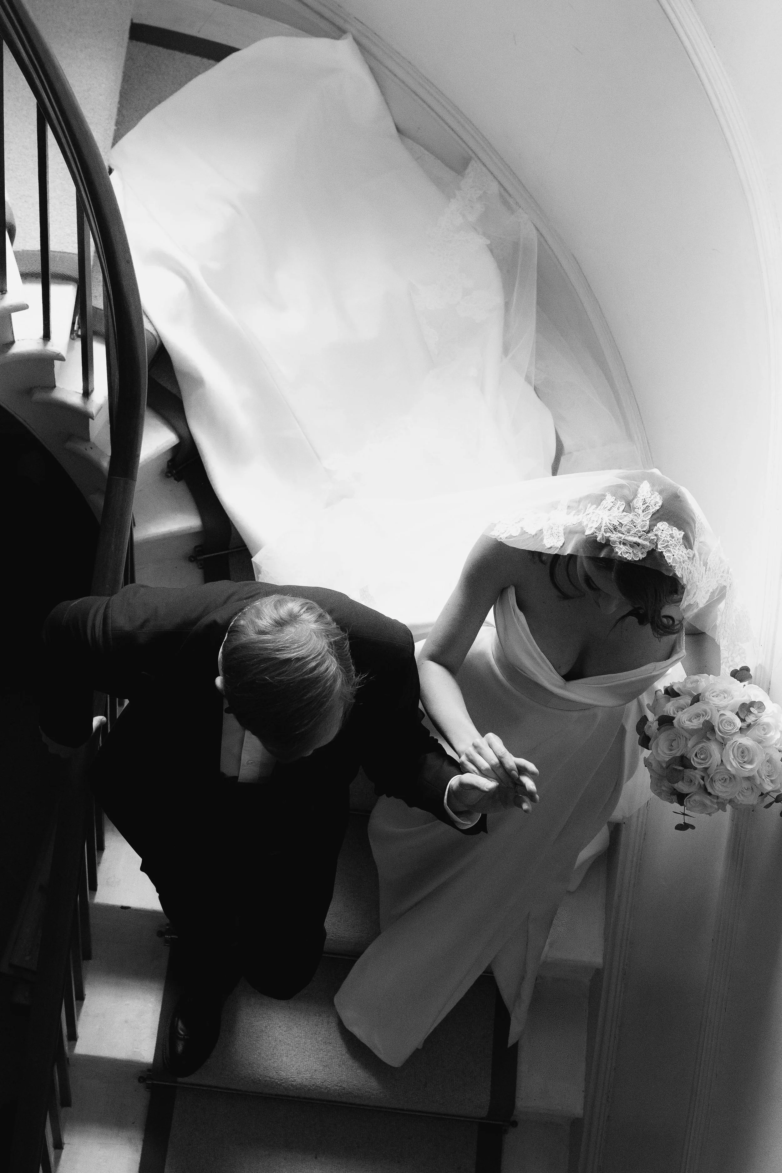 A bride in a wedding gown holding a bouquet of roses is praying with a groom in a suit in a small, well-lit room, possibly in a hotel or chapel.
