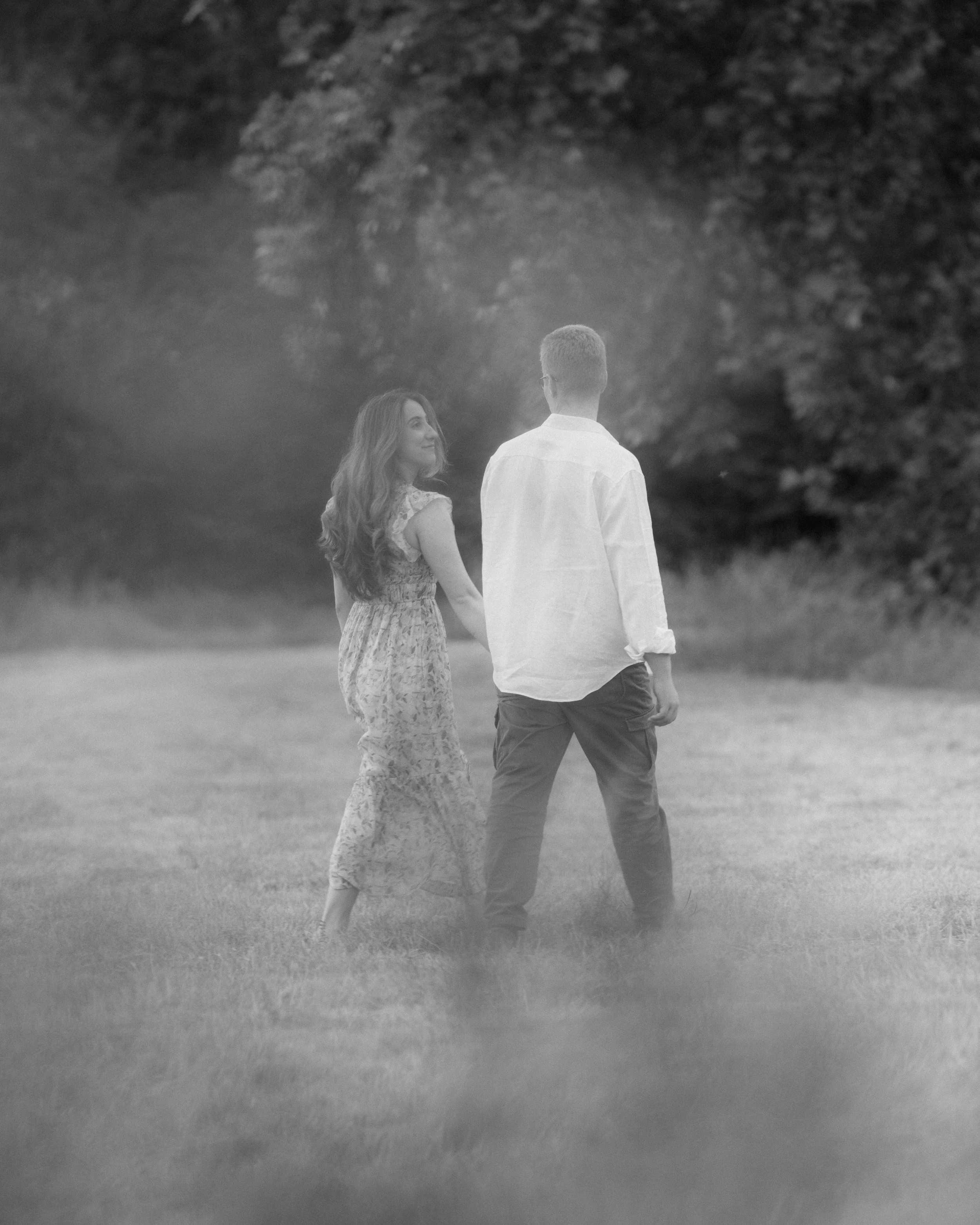 A man and woman holding hands and walking outdoors in a grassy area near trees, black and white photograph.