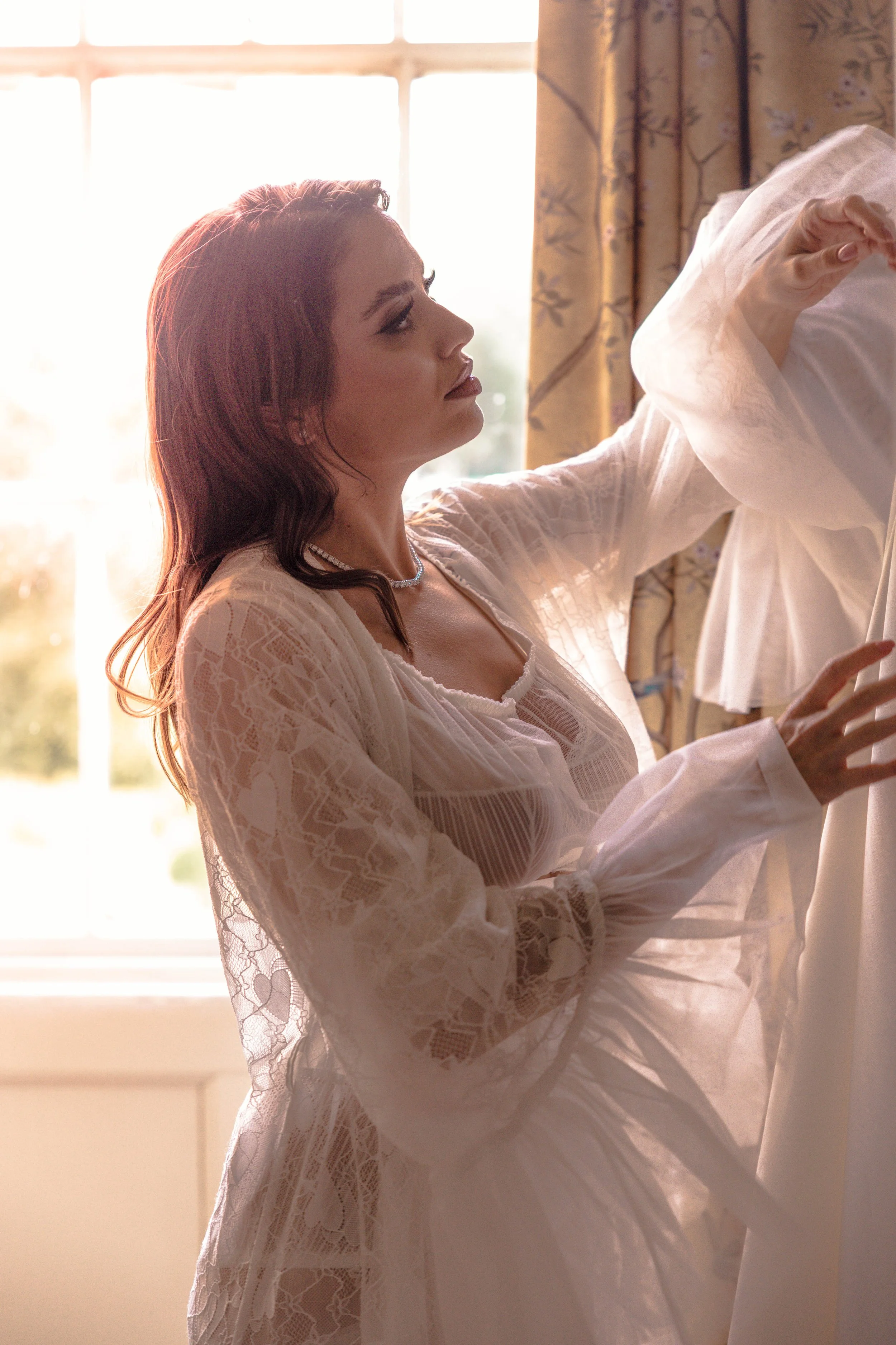 A woman with long, wavy brown hair and makeup, wearing a white lace robe, sitting by a window in soft sunlight, adjusting her wardrobe.