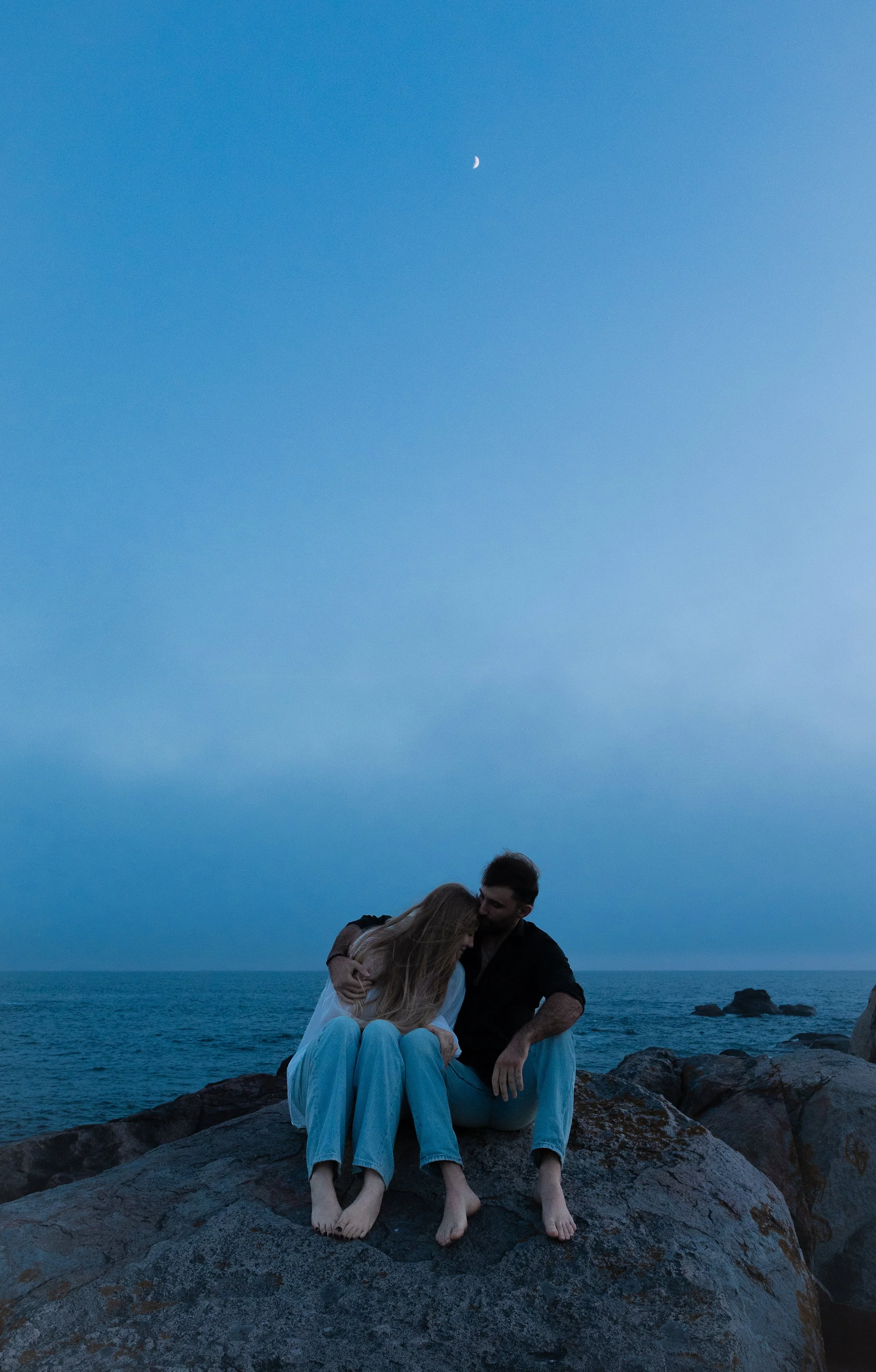 A couple sitting on rocks by the ocean at dusk, embracing and sharing an intimate moment, with the moon visible in the sky.