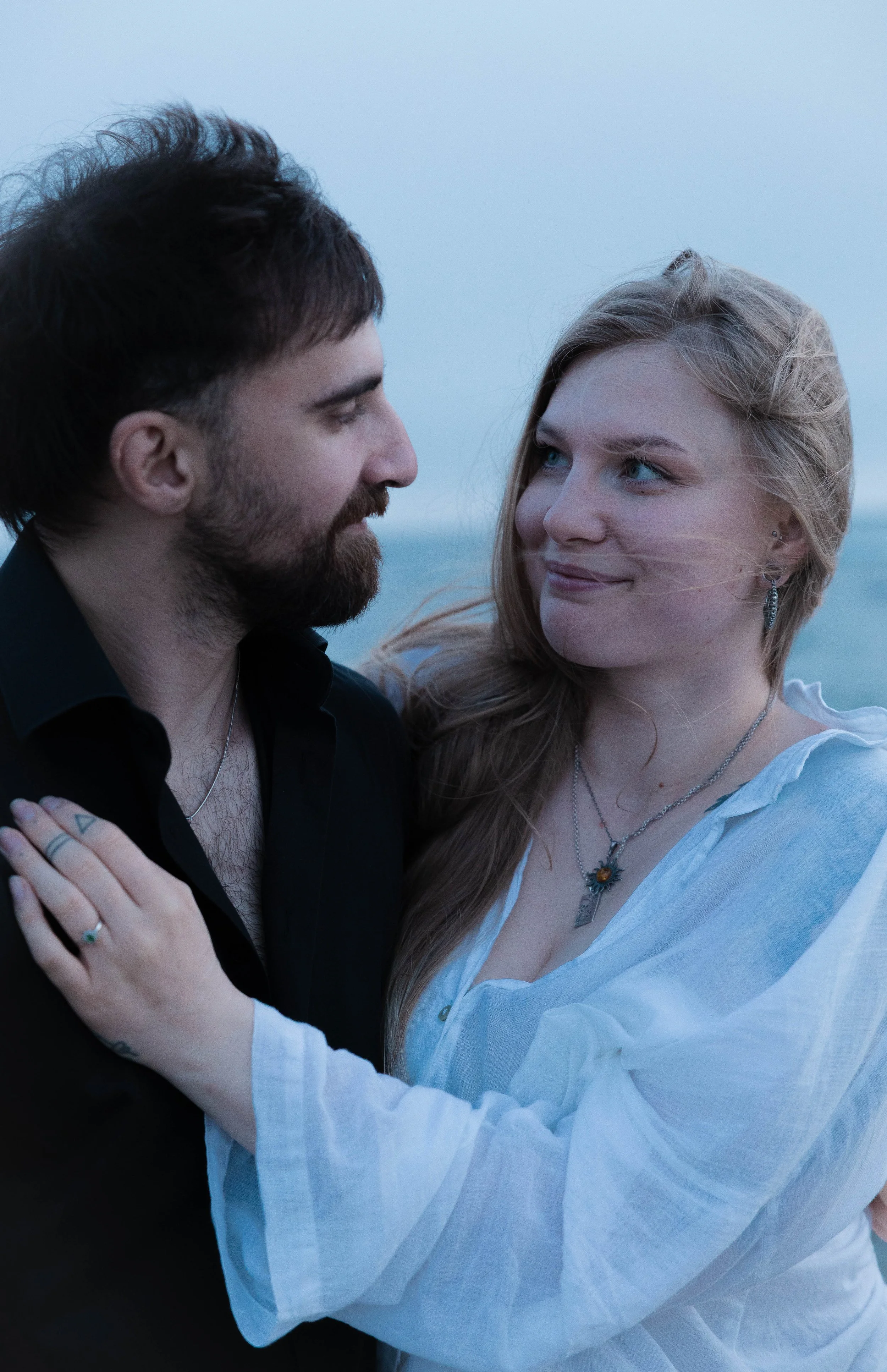 A man and woman facing each other closely, holding each other by the shoulders, outdoors near water, with a light sky in the background.