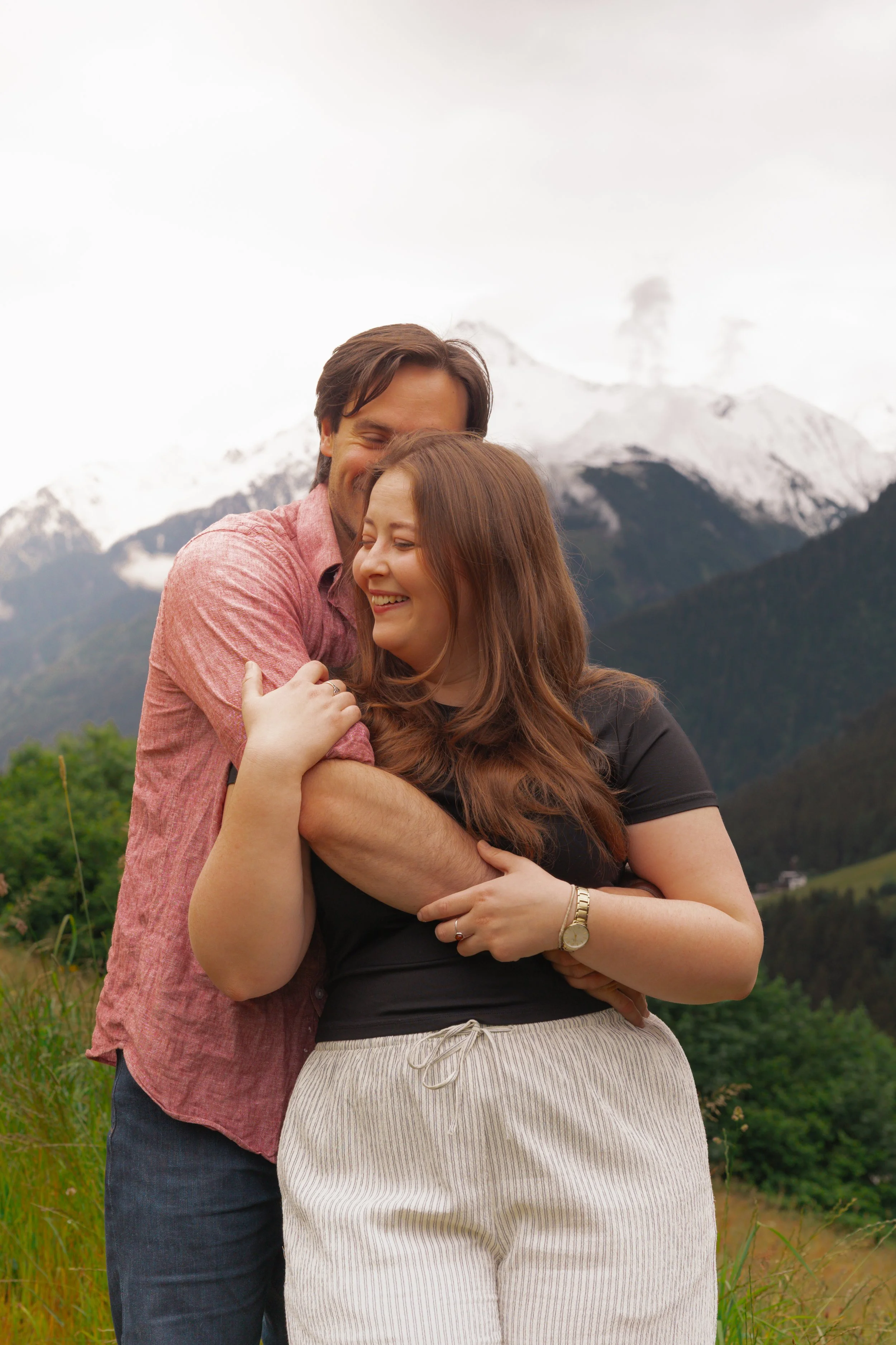 A man and a woman hugging outdoors with snow-capped mountains in the background.