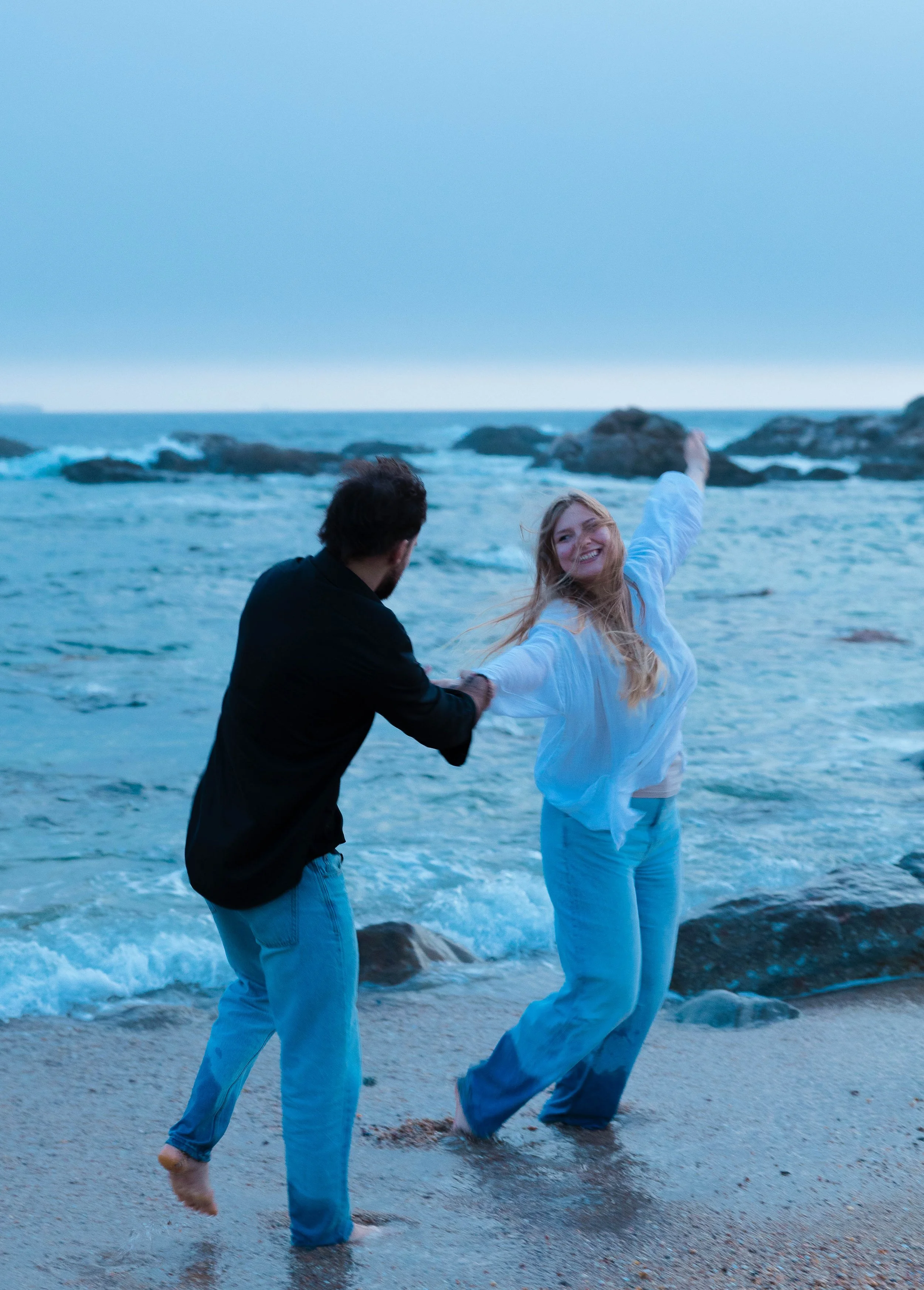 Two people, a man and a woman, are standing ankle-deep in the water on a beach. The man is facing away and wearing a black jacket and blue jeans. The woman, smiling and holding her arms out, is wearing a white shirt and blue jeans. The ocean waves and rocks are visible in the background, with a cloudy sky overhead.