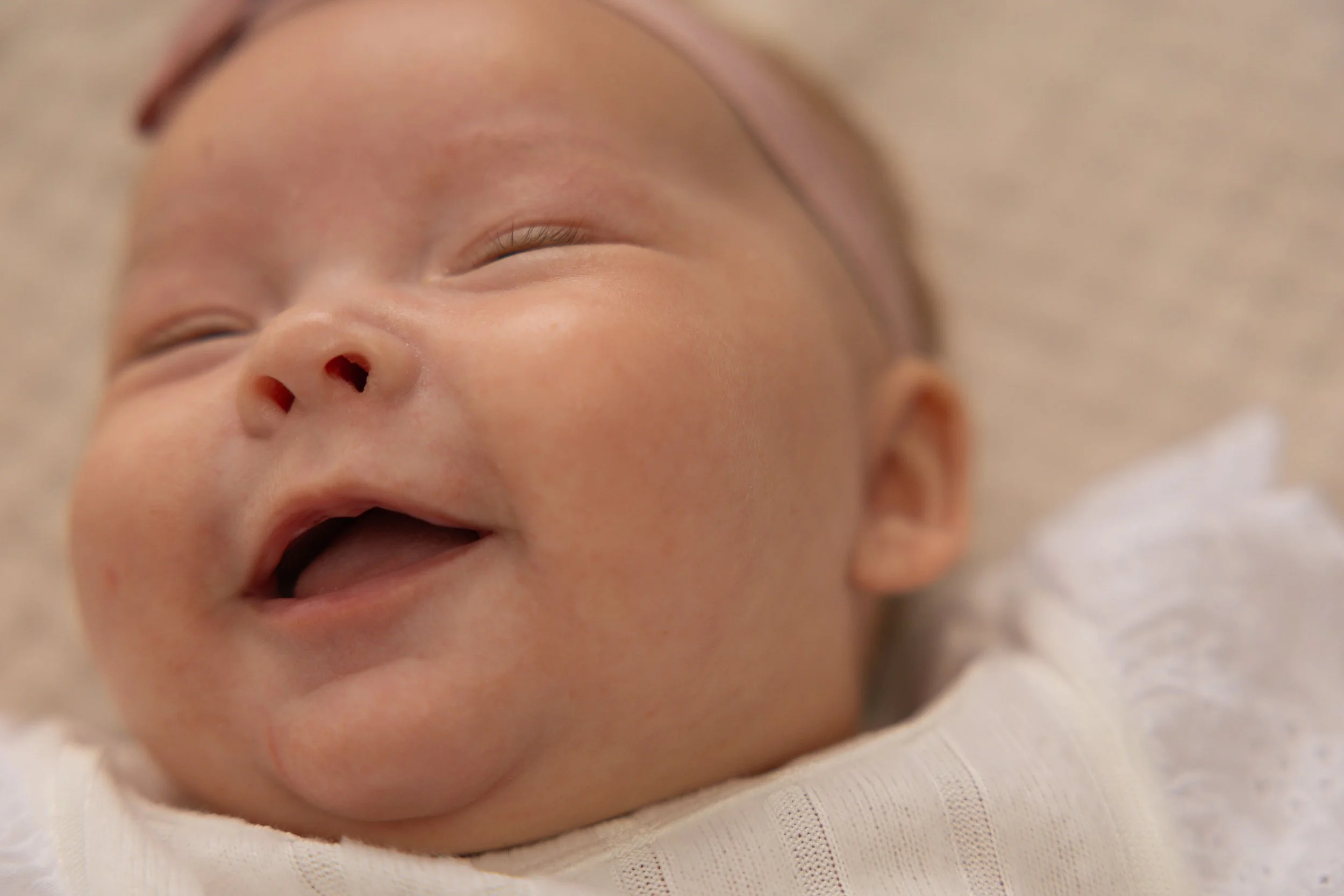 Close-up of a smiling baby's face with eyes closed and mouth open, lying on a soft surface.