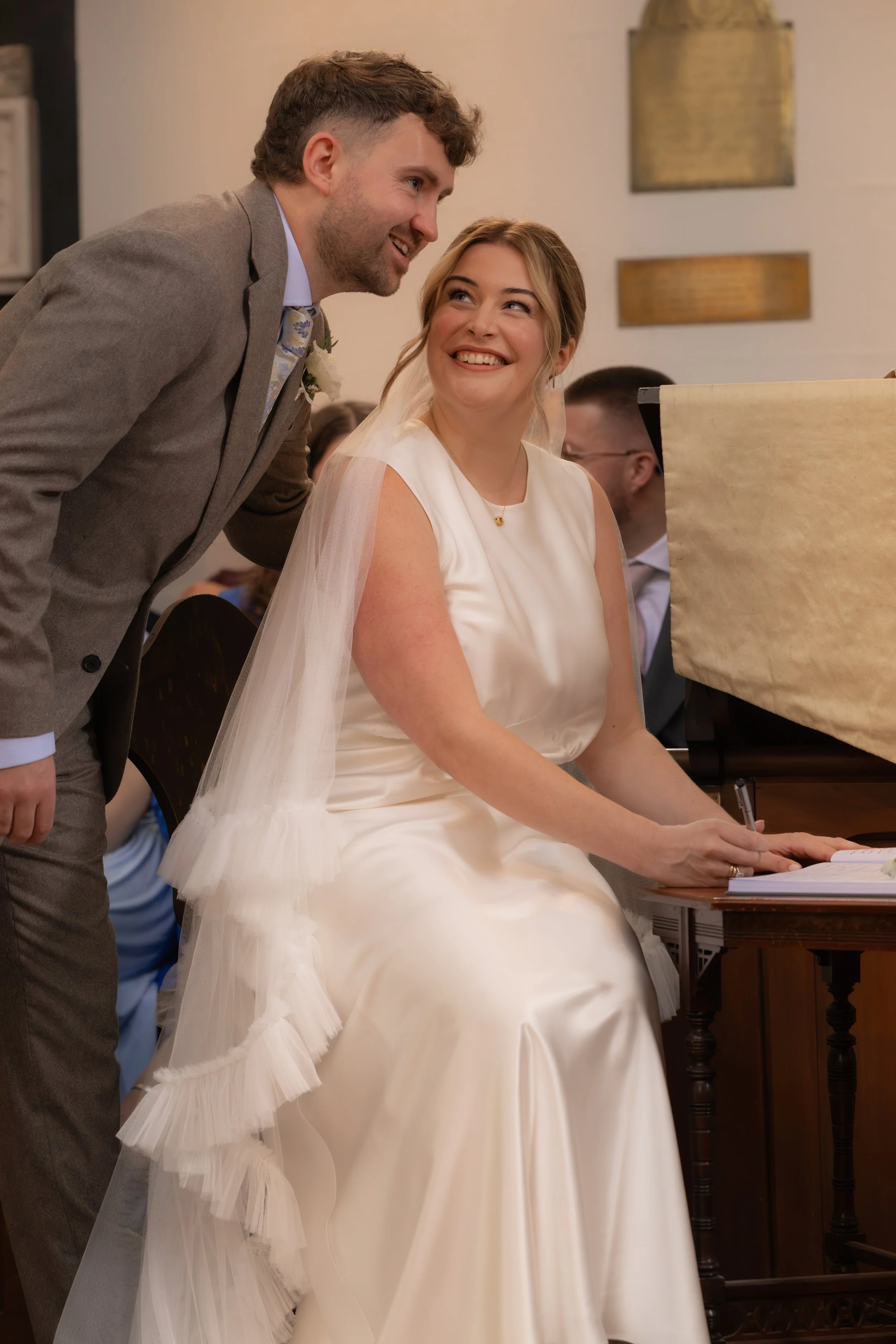 A bride in a white wedding dress sitting at a table, smiling as a man in a gray suit leans toward her, at a wedding.