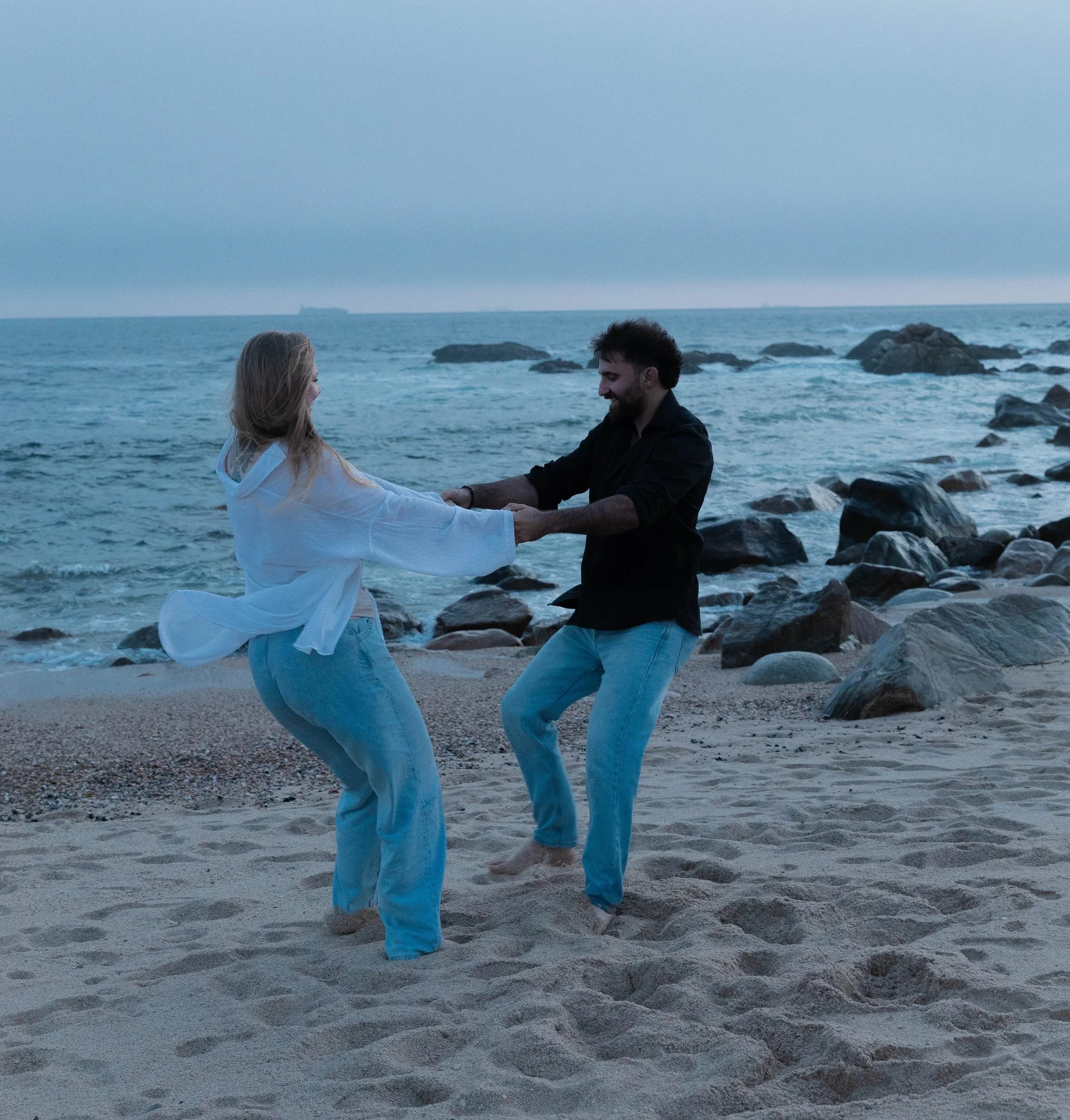 A couple holding hands and facing each other on a beach with rocks and the ocean in the background, with ship in the distant horizon, during evening or dusk.