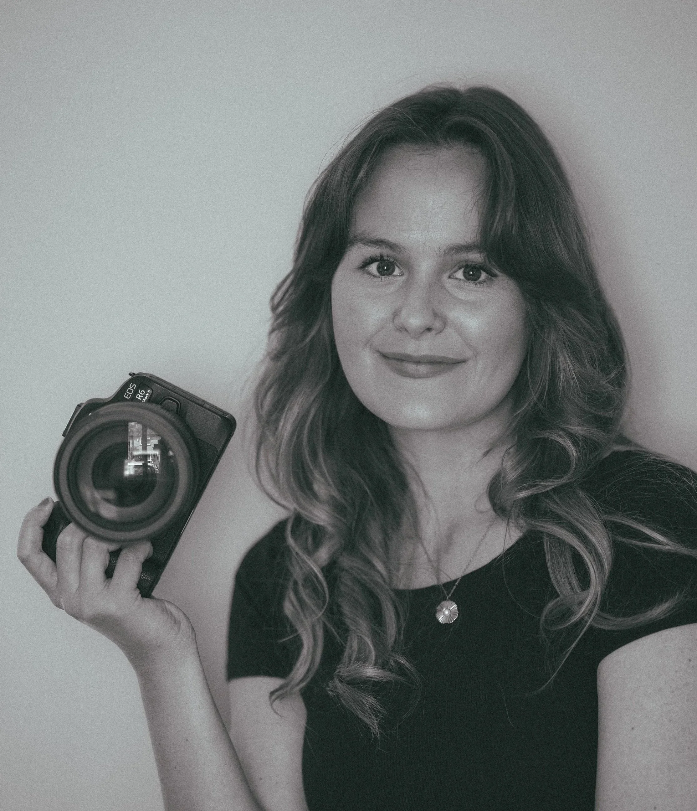 Black and white photo of a woman with long, wavy hair holding a camera, taking a selfie, with part of her face visible and wearing a top with a round brooch.