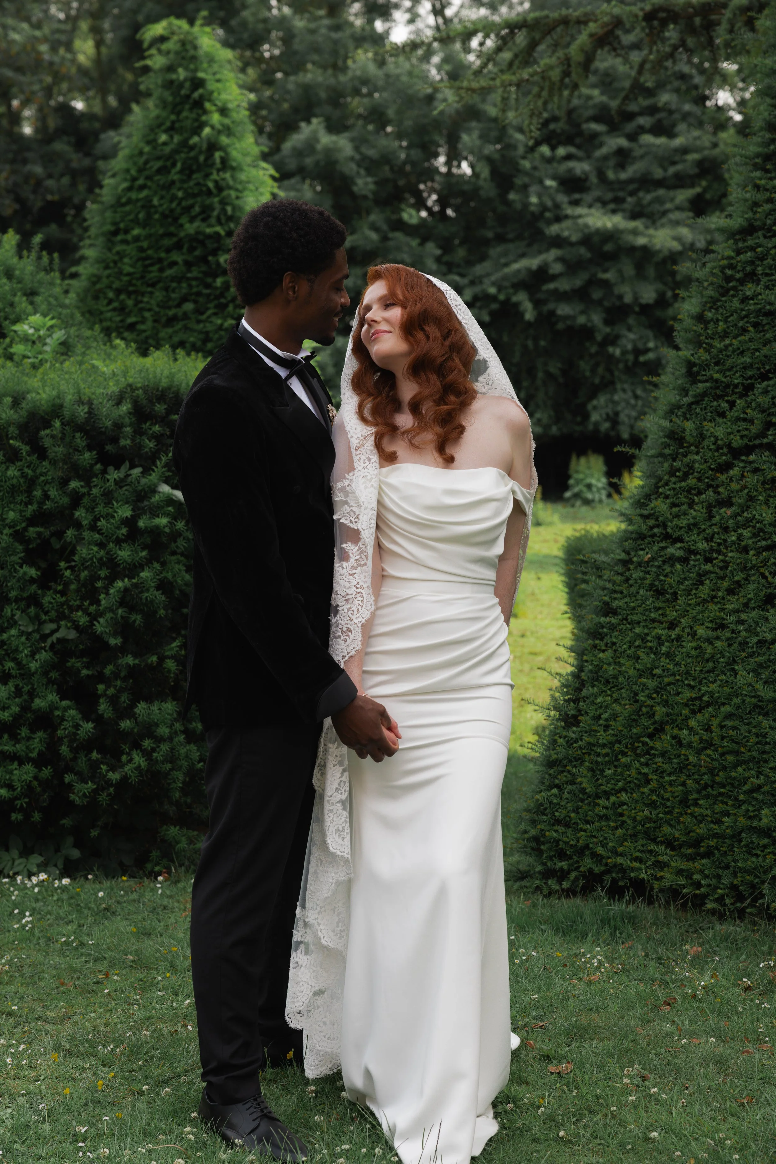 A bride and groom holding hands and leaning towards each other in an outdoor garden setting, with trees and greenery in the background.