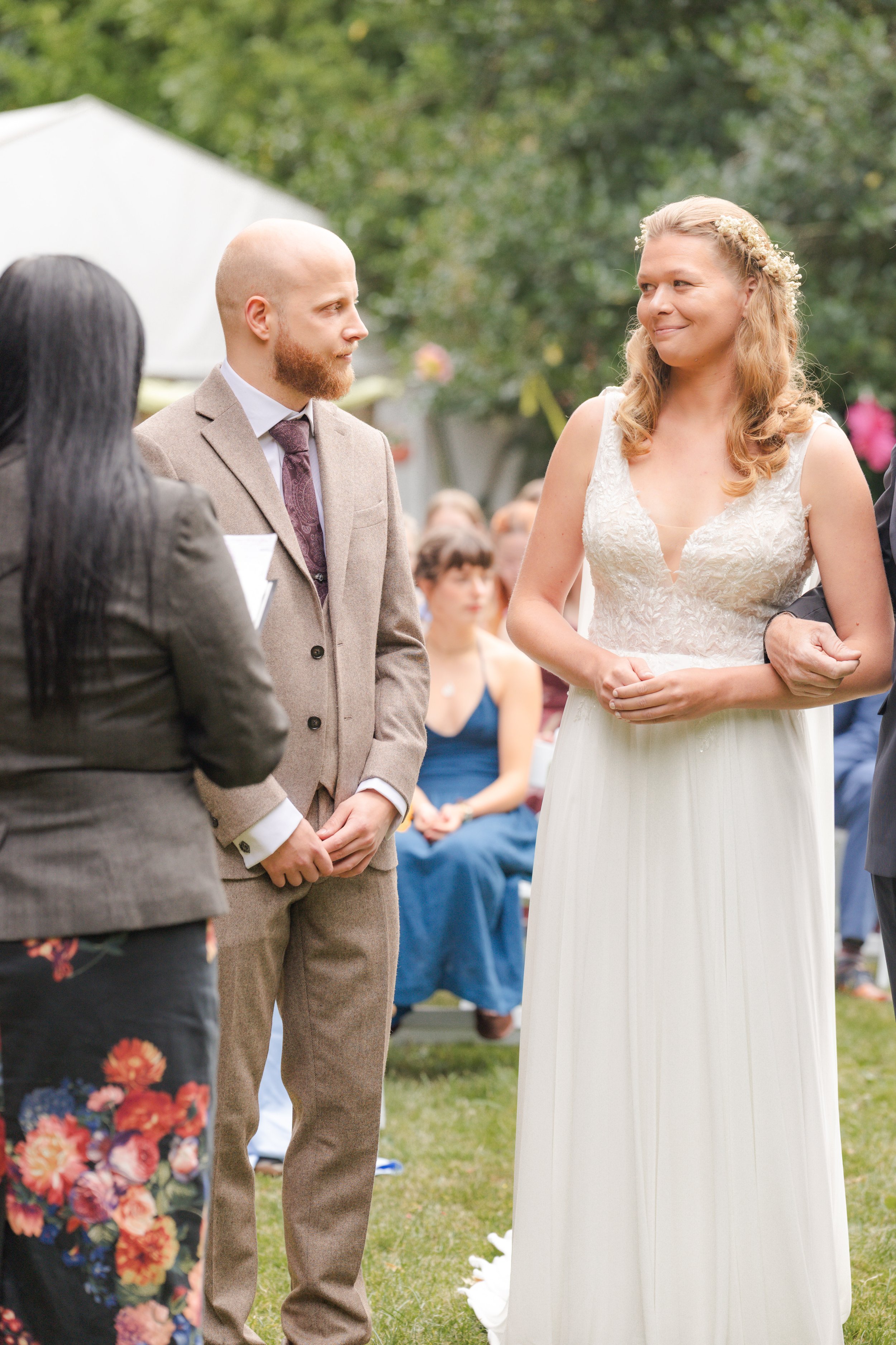 A couple exchanging vows at a wedding ceremony outdoors with guests seated in the background. The bride wears a white lace dress with a floral crown, and the groom wears a beige suit with a tie.