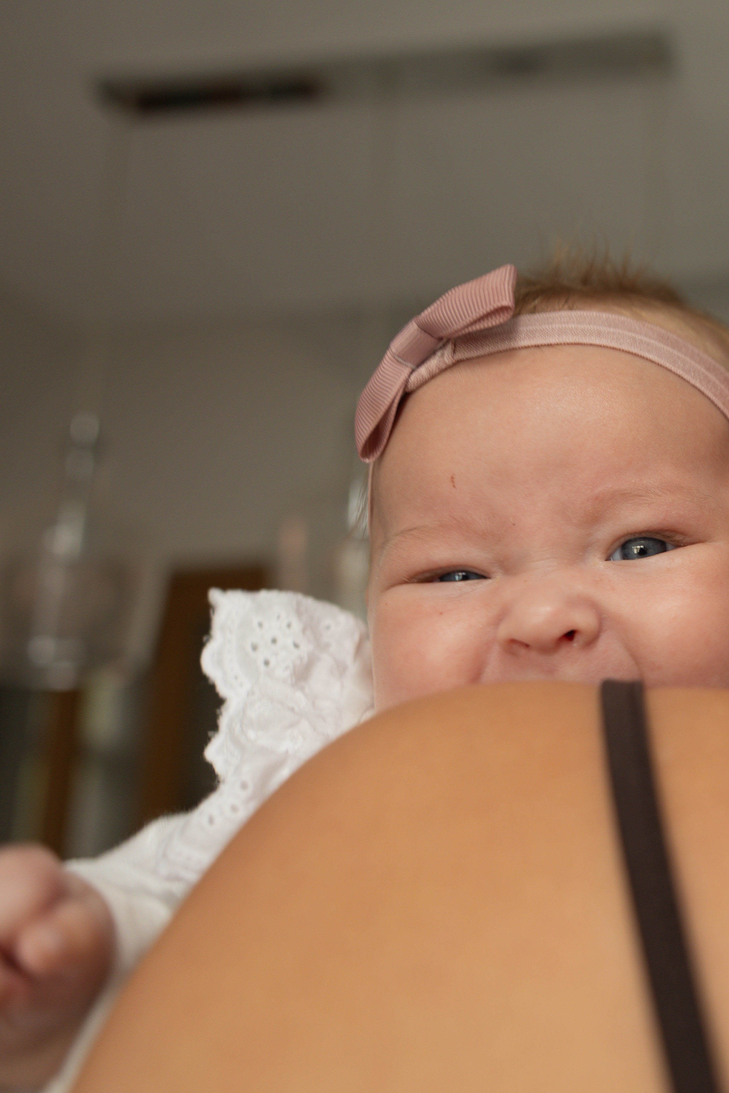 Close-up of a smiling baby girl wearing a pink headband with a bow, a white lace sleeve, and a black strap, with a blurred background.