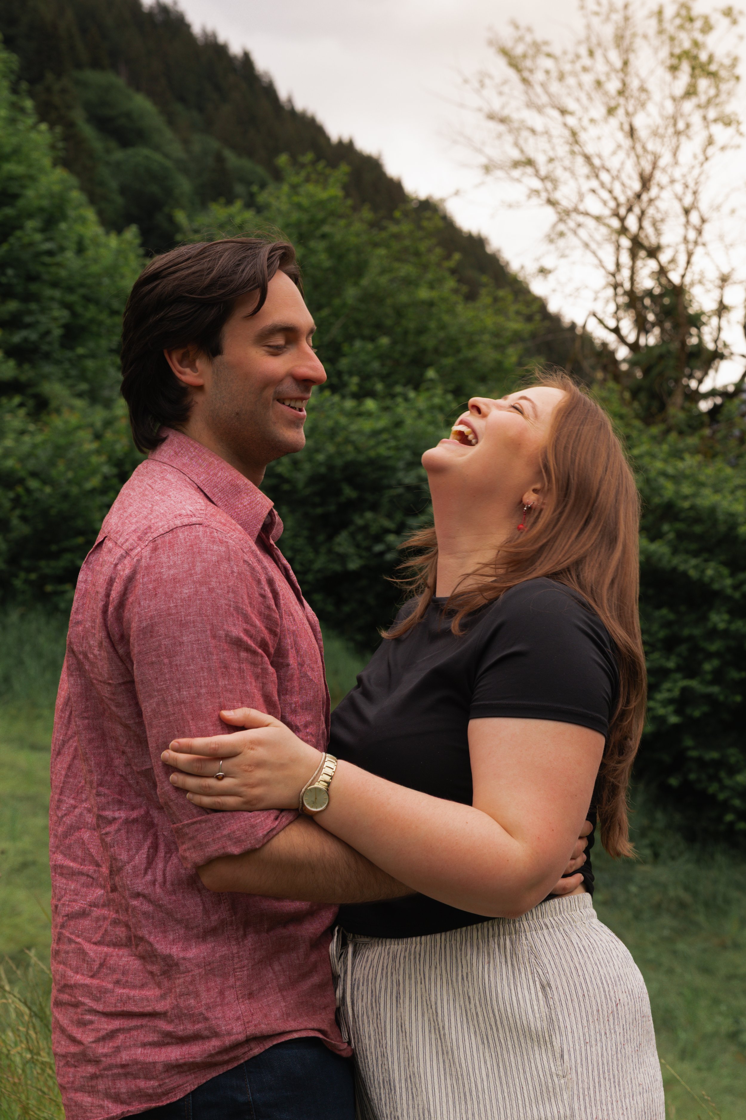A happy couple standing outdoors in a green, wooded area. The man has brown hair and is wearing a pink shirt, and the woman has red hair and is wearing a black top and striped skirt. They are smiling and looking at each other, embracing with the woman's hand on the man's arm.