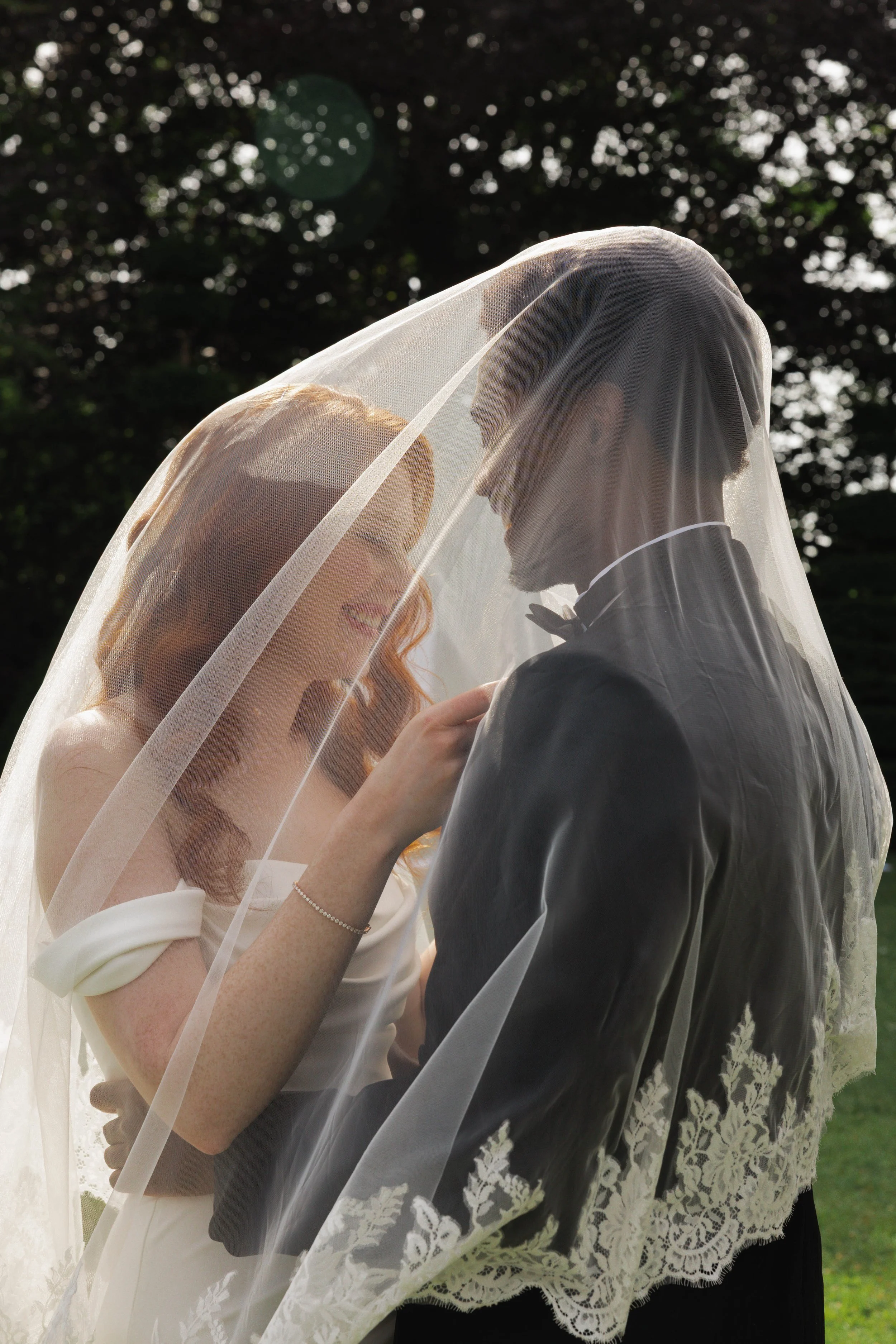 A bride and groom sharing a moment under a veil outdoors, with trees in the background.