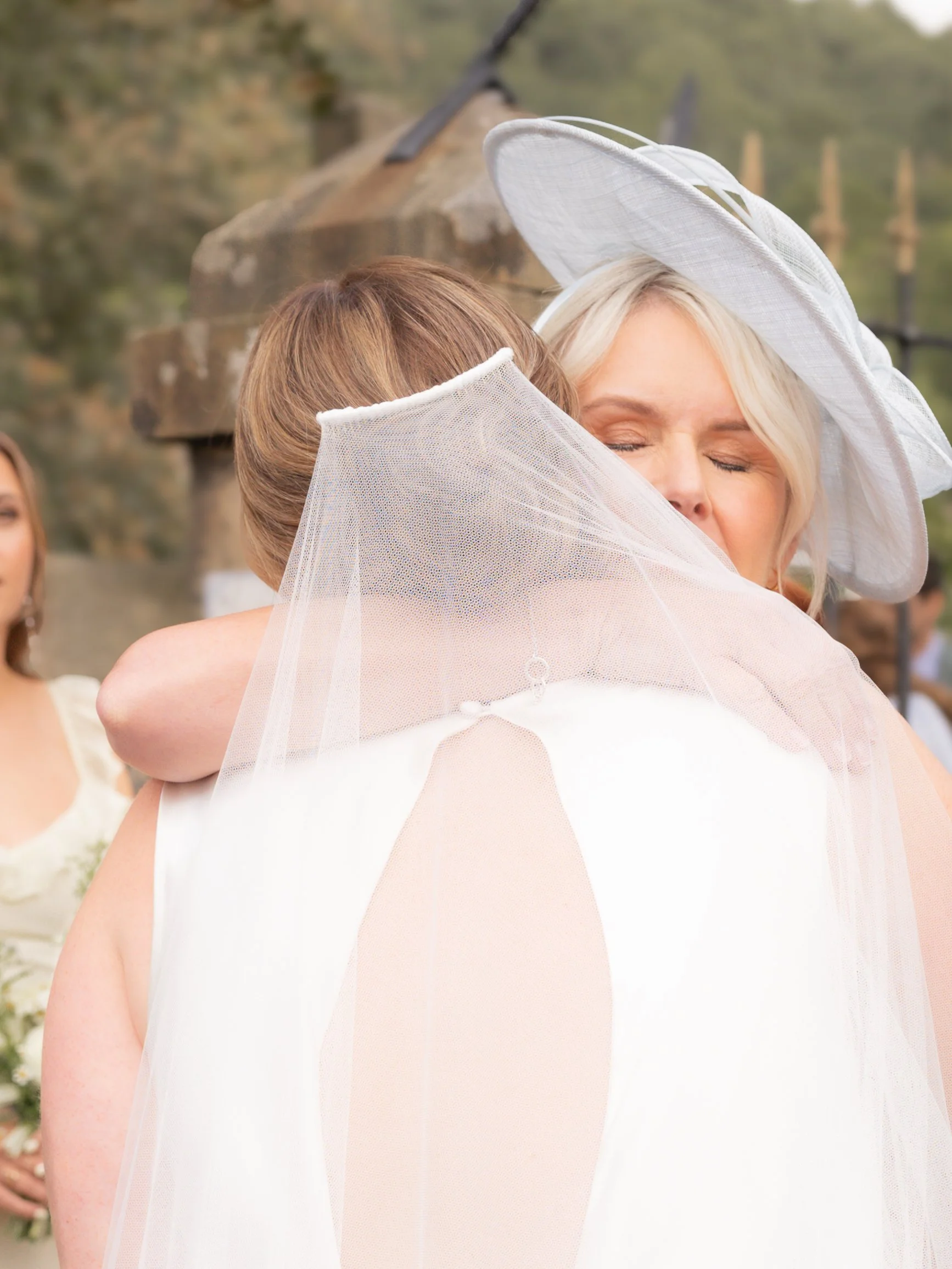 Bride and her mom hugging after the wedding ceremony, outdoors with trees in the background.