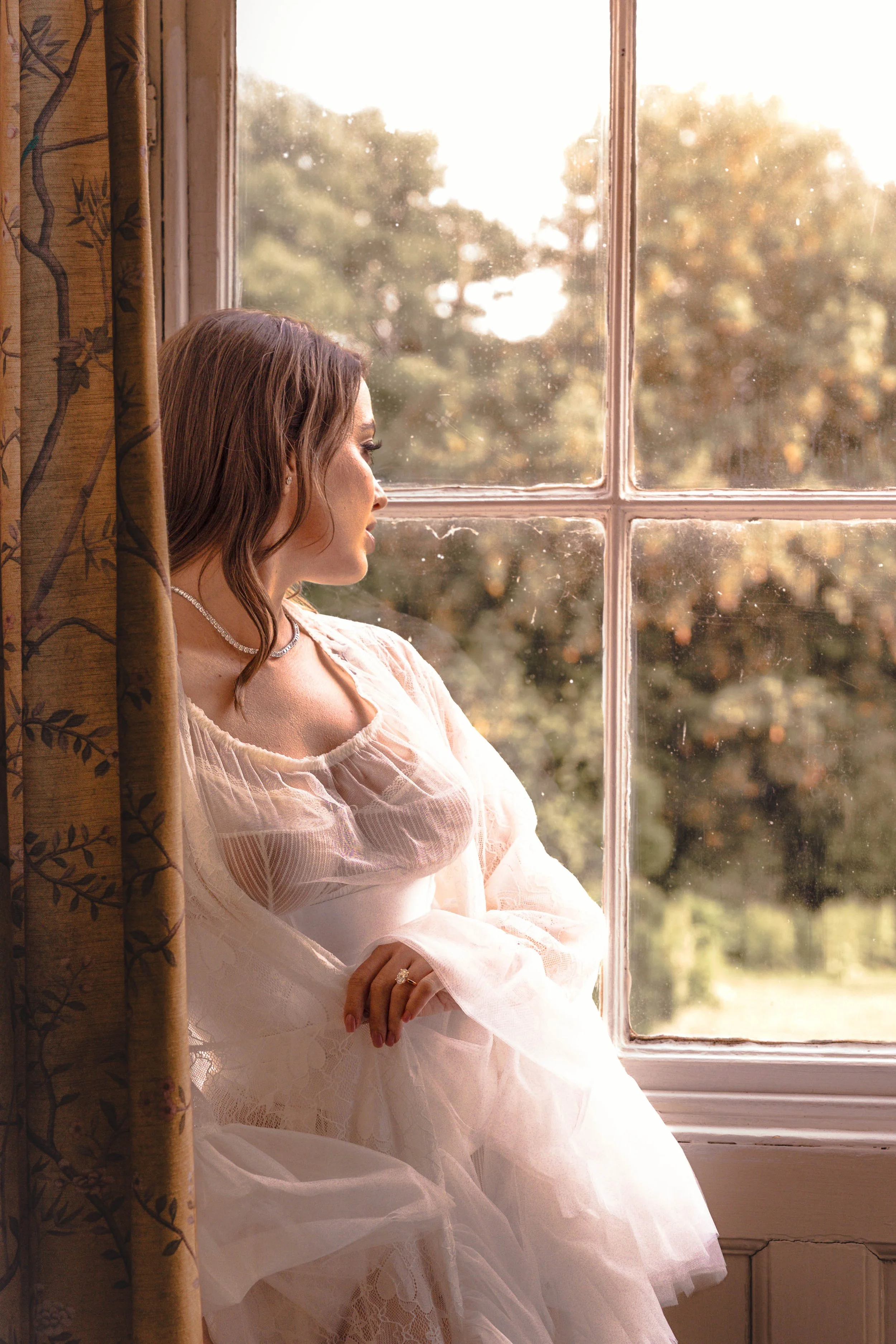 A woman with wavy brown hair in a white, lace dress, wearing a pearl necklace and ring, looking out a window with a view of trees and sunlight, partially obscured by floral curtains.