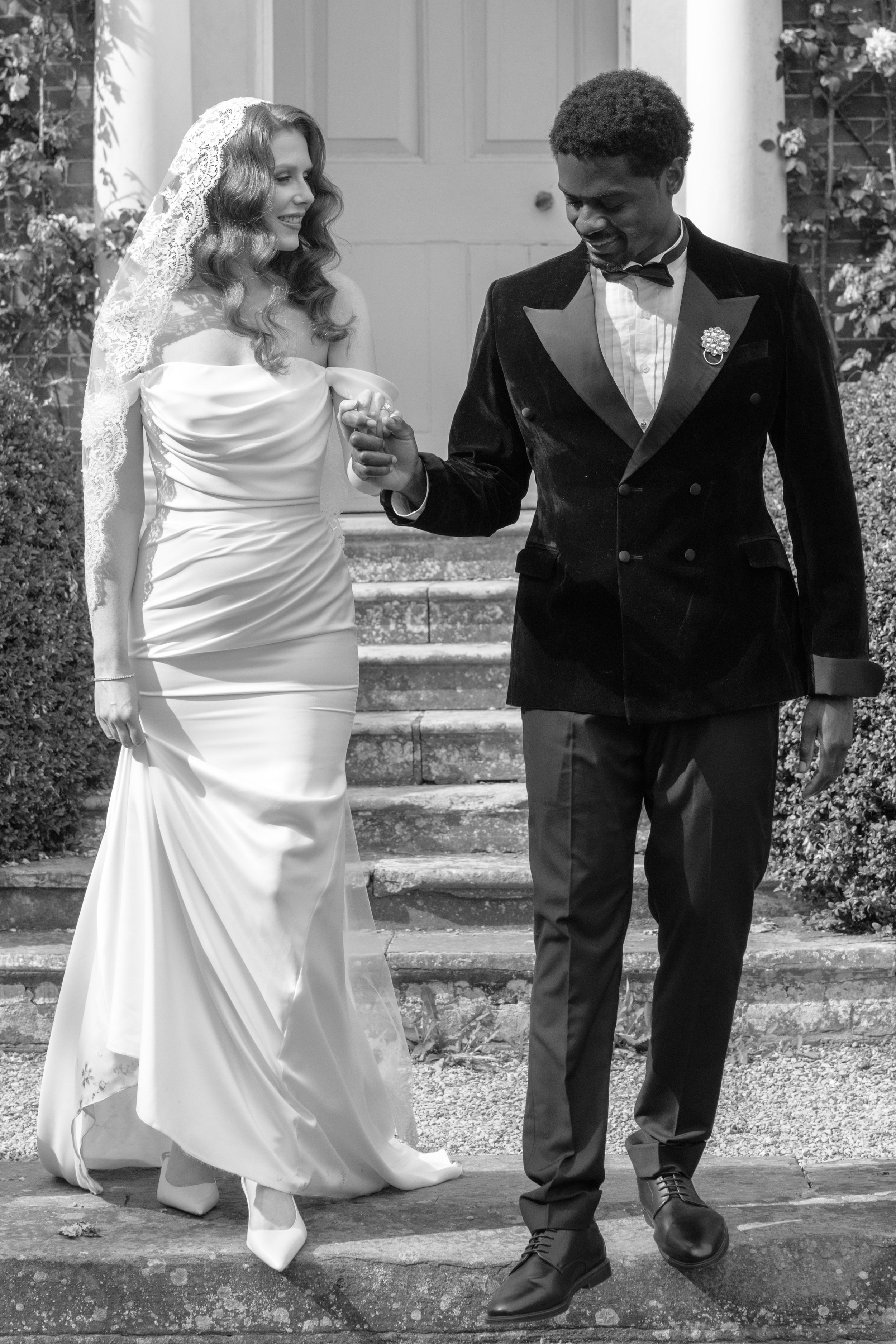 A black and white photo of a bride and groom smiling and holding hands while walking down the steps outside a house. The bride is wearing a strapless wedding gown with lace details and a veil, and the groom is dressed in a tuxedo with a bow tie and a