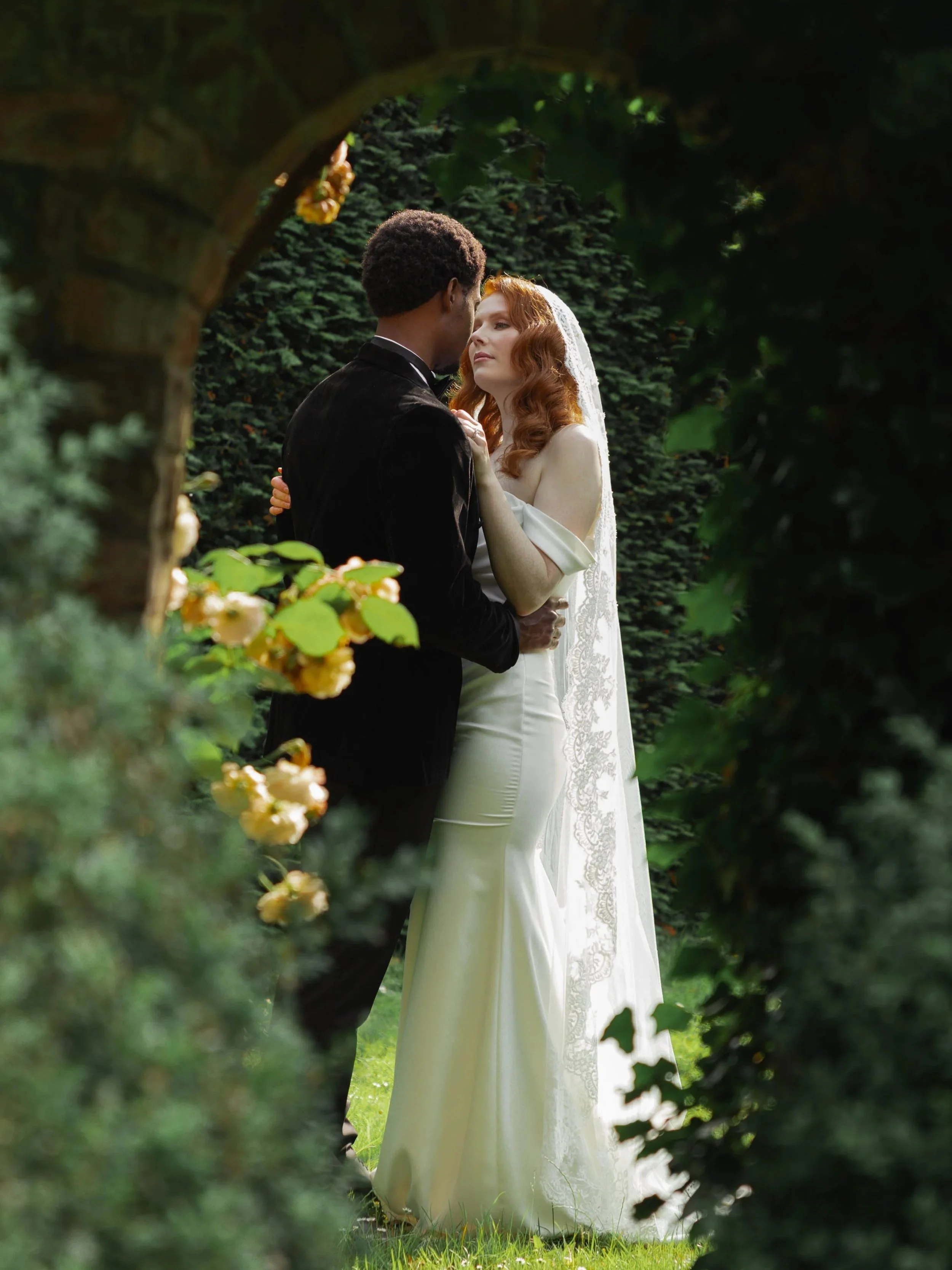 A bride and groom are standing close together outdoors, framed by a stone archway and surrounded by greenery, as they gaze into each other's eyes.