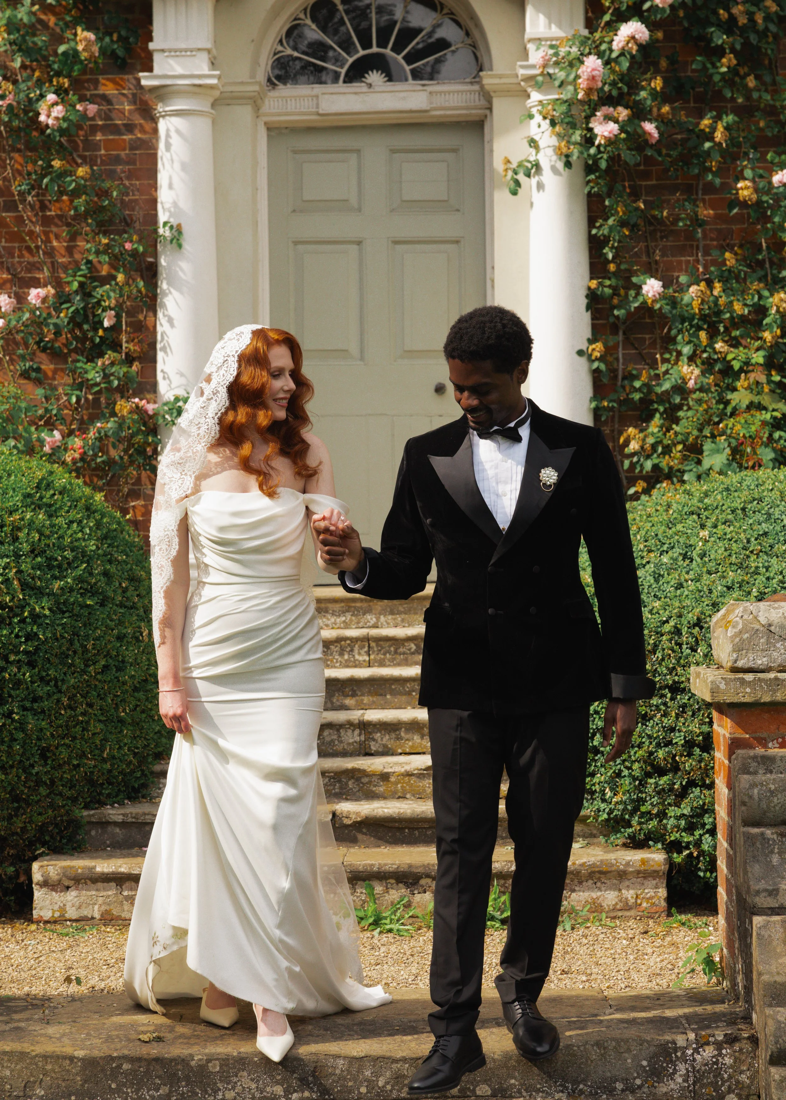 A bride with red hair in a white wedding dress and a groom with dark skin in a black tuxedo are walking up stairs outside a brick house covered in flowering vine, holding hands and looking at each other.