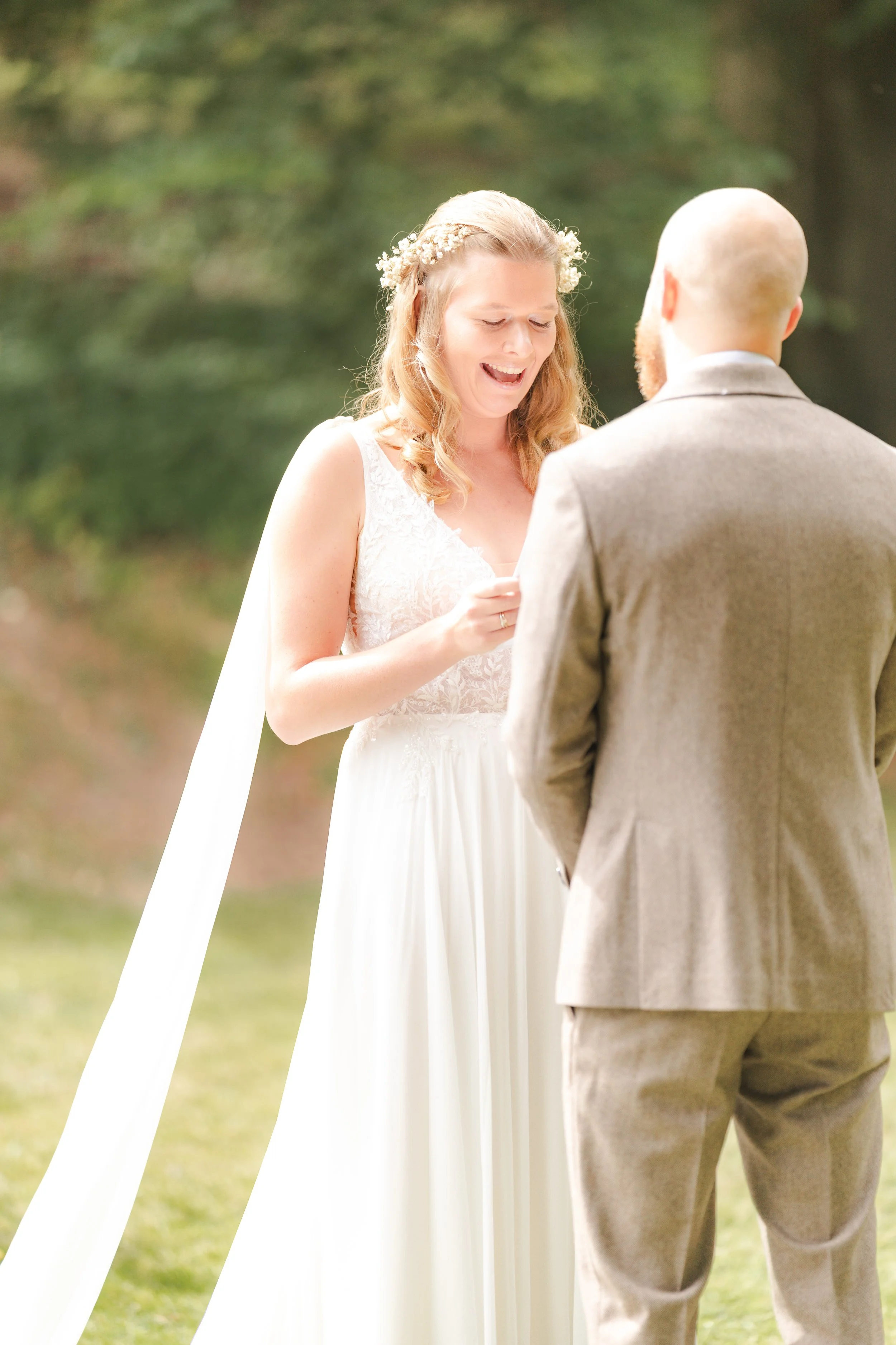 A bride with light brown hair and a floral headband is smiling as she looks down at her hand during a wedding ceremony outdoors. She is wearing a white sleeveless wedding dress. A man with a bald head and beard, dressed in a gray suit, stands in fron