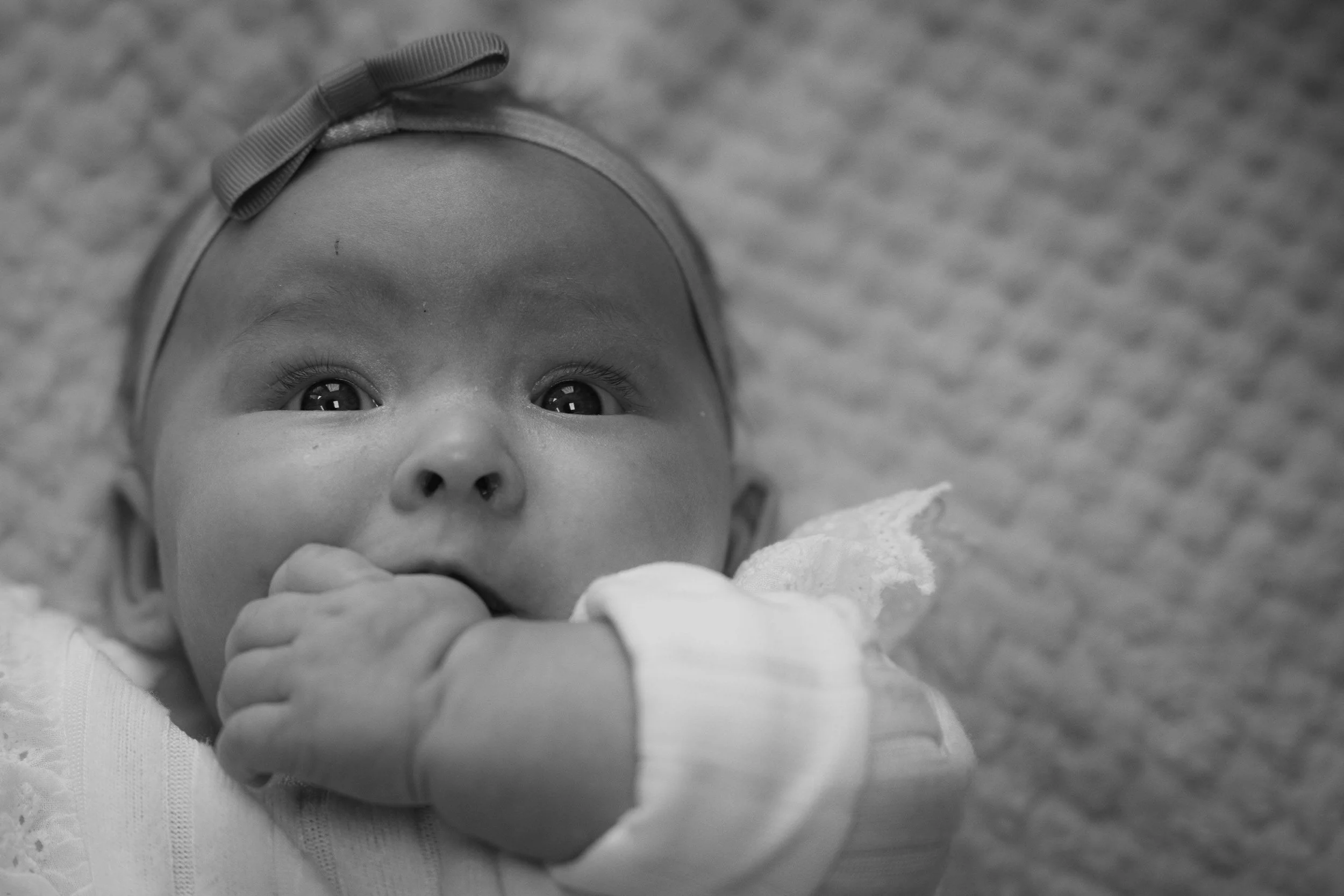 A close-up black-and-white photo of a young child looking upward with curious eyes, holding a soft object near their mouth, wearing a headband with a bow, and lying on a textured surface.