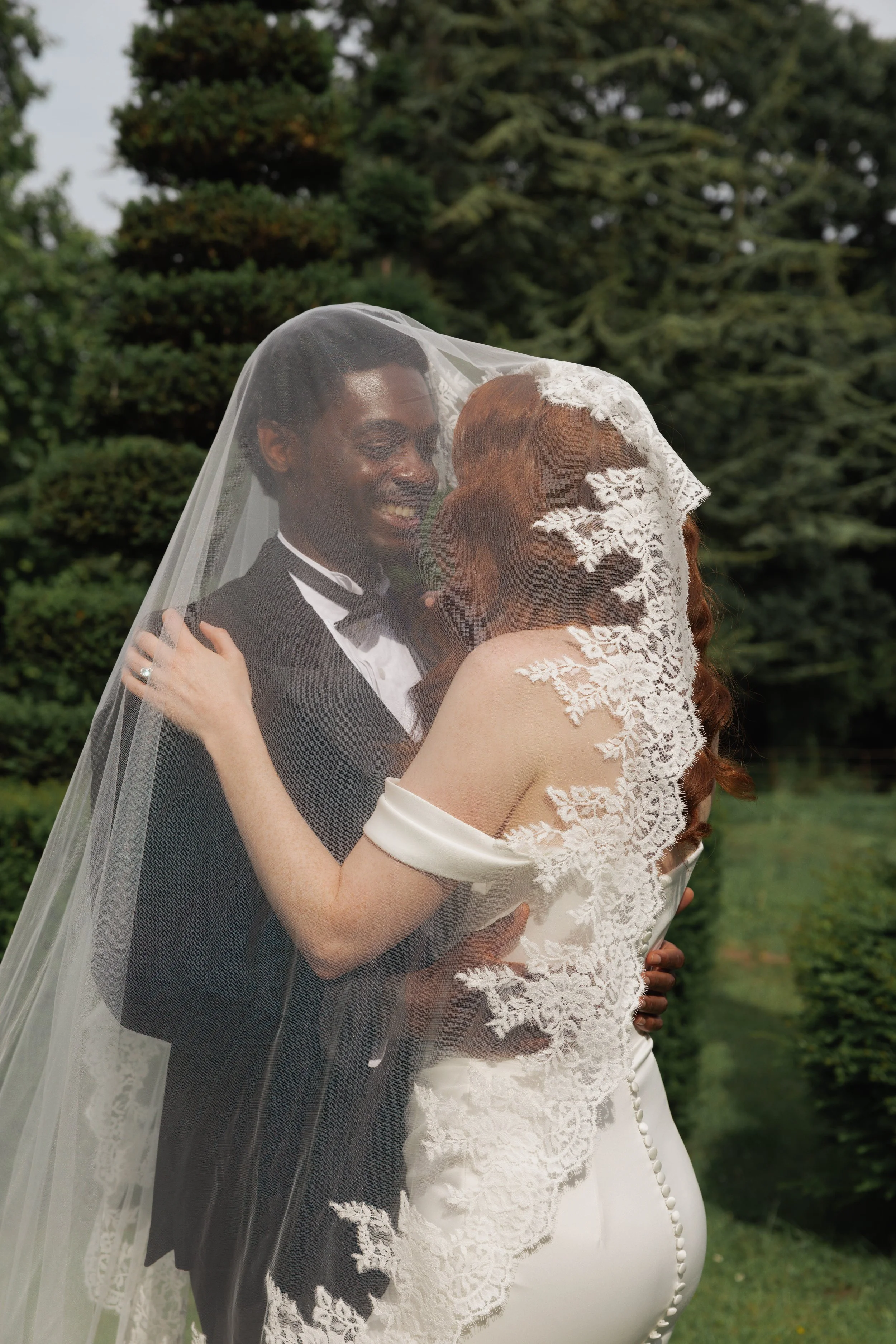 Couple dressed in wedding attire, smiling and embracing outdoors under a leafy tree, with a bride wearing a lace wedding dress and veil, and a groom in a tuxedo.